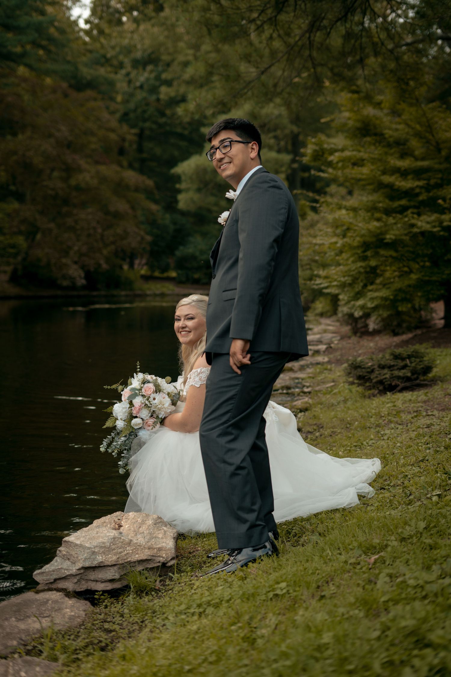 Bride seated on rocks holding pink bouquet with groom standing beside a serene woodland lake.