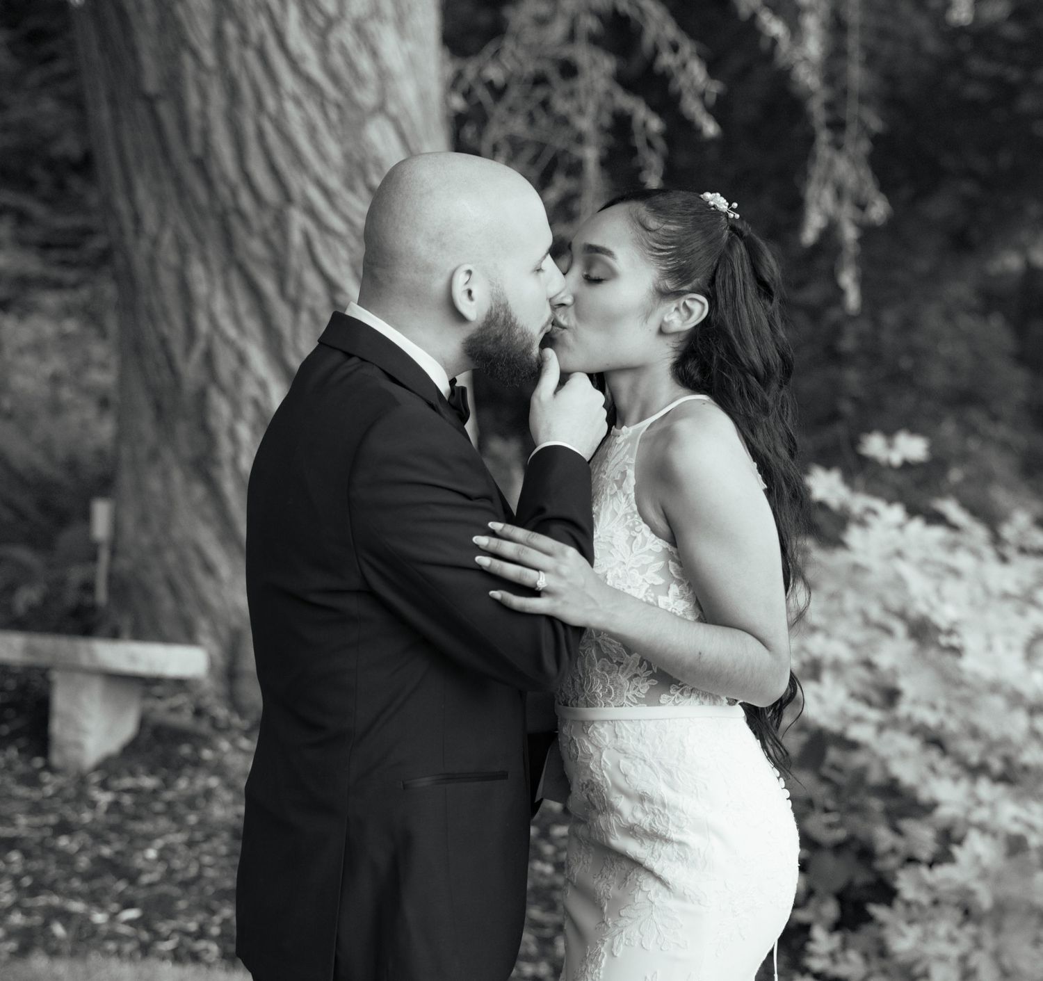 Black and white photo of bride and groom sharing a tender kiss among trees on their wedding day.