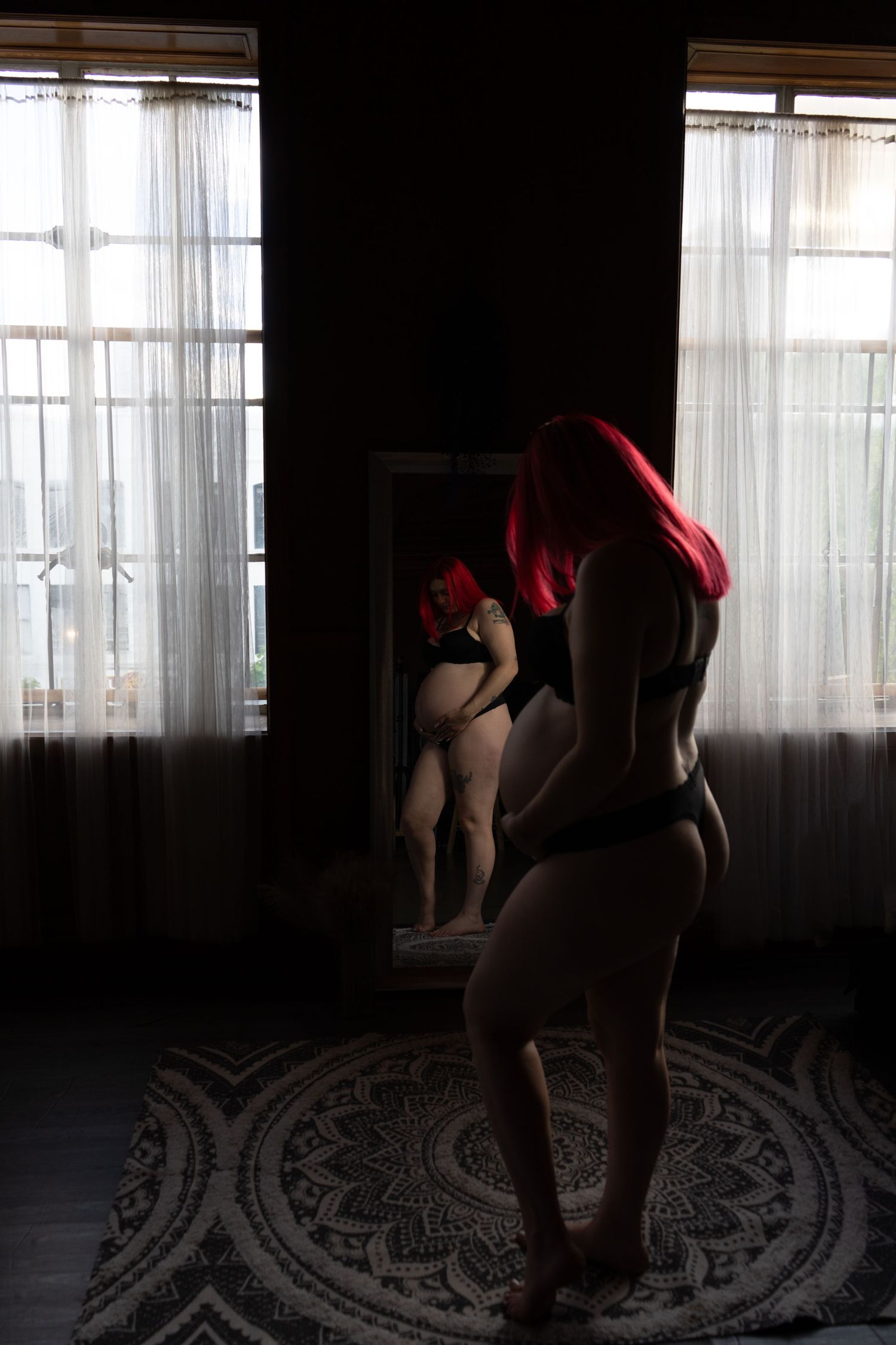 Red-haired woman in black lingerie gazes at reflection in mirror in dramatic dark room.