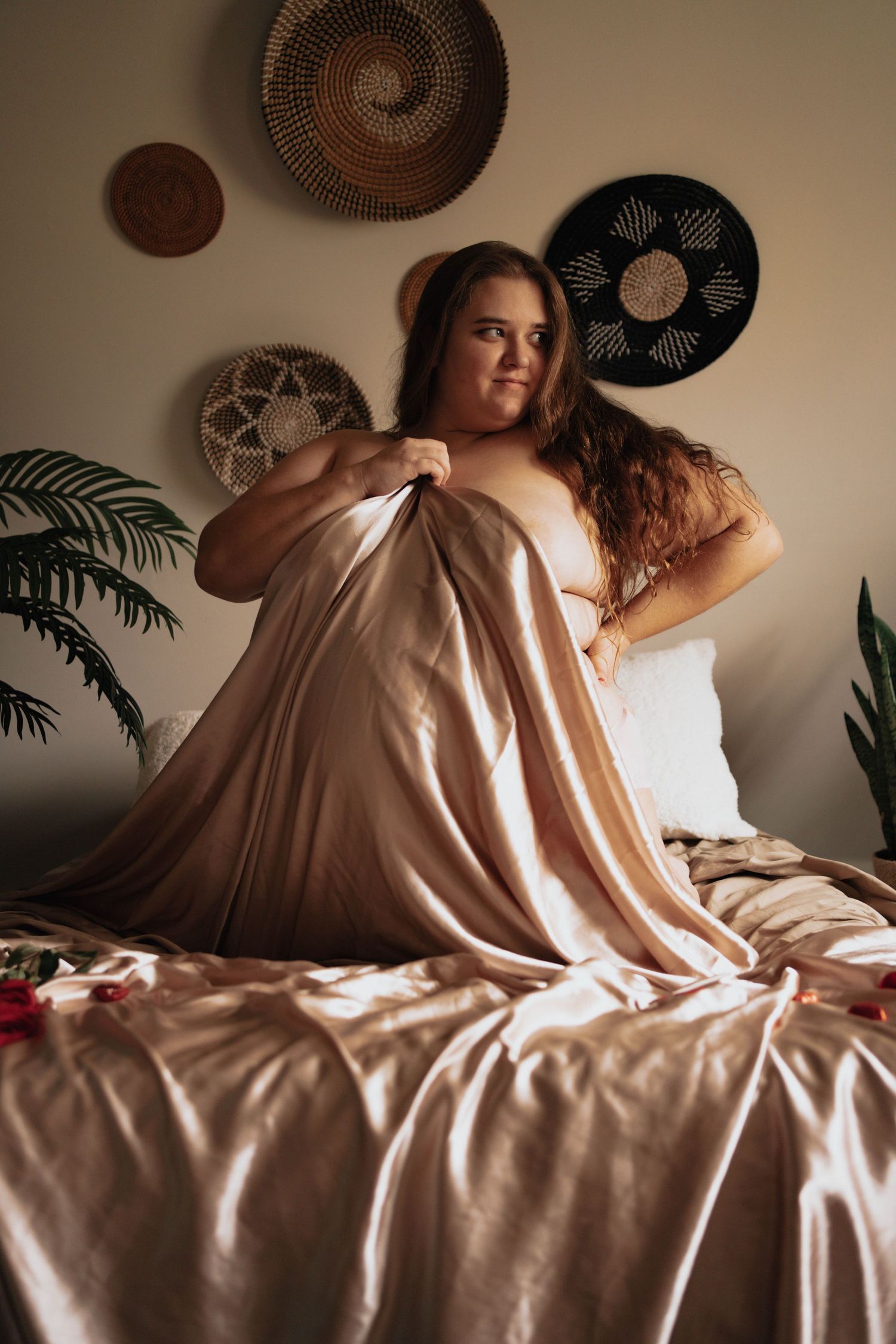 A plus-size woman sitting on a bed wrapped in luxurious satin sheets in a boho-decorated bedroom.