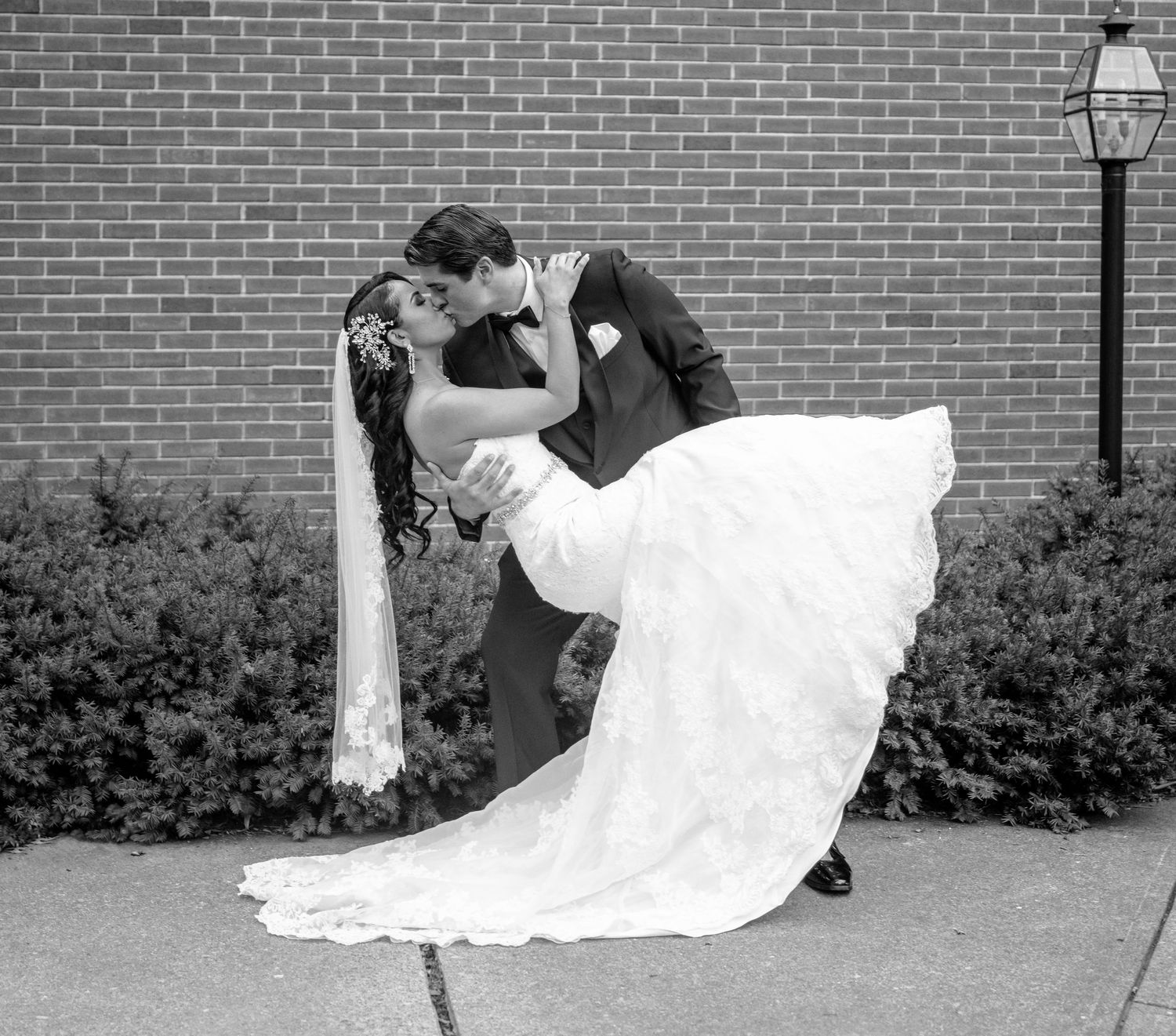 Black and white photo of groom dipping bride in a lace wedding gown outside a brick building.