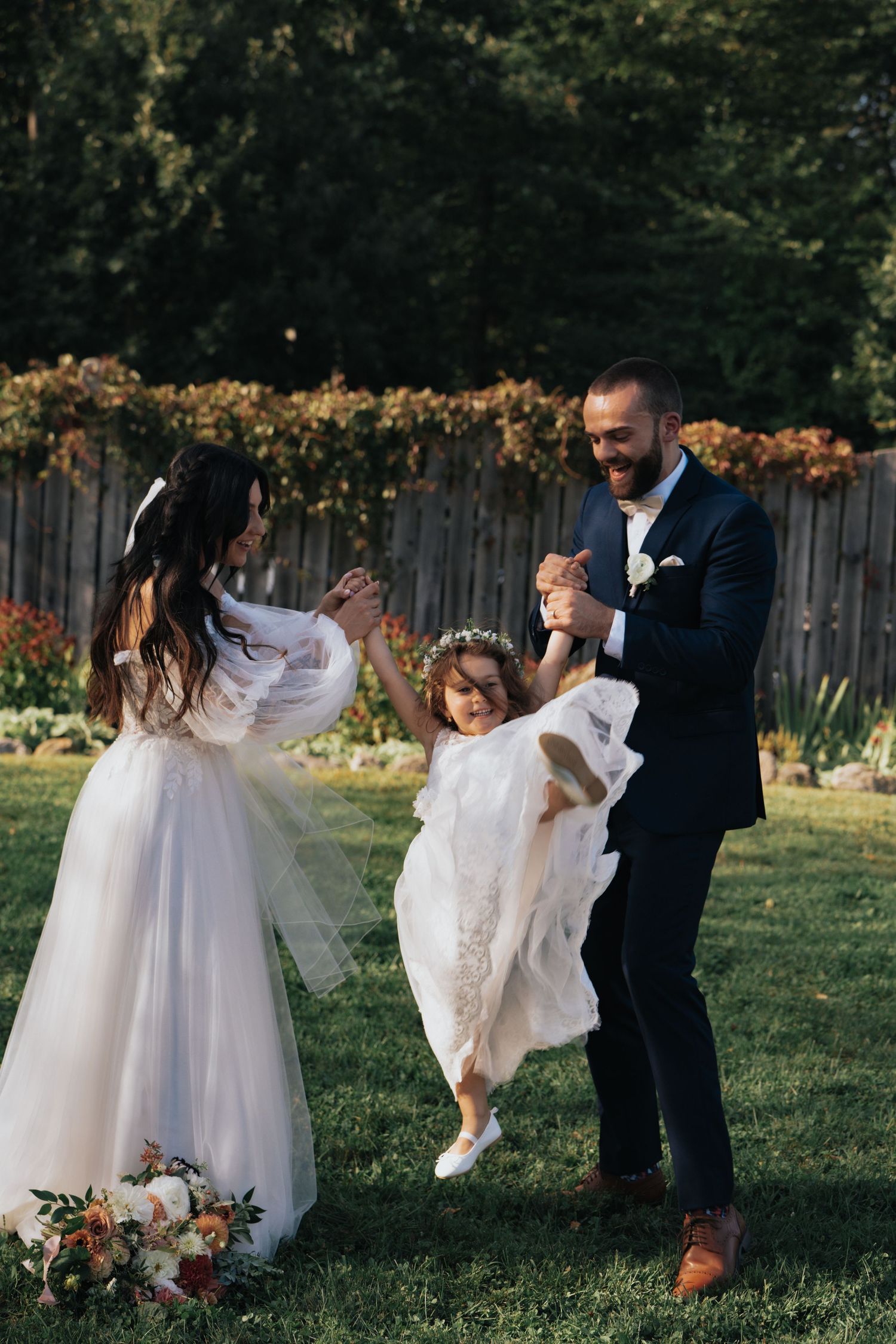 Bride and groom joyfully swinging flower girl between them during outdoor wedding celebration on green lawn.