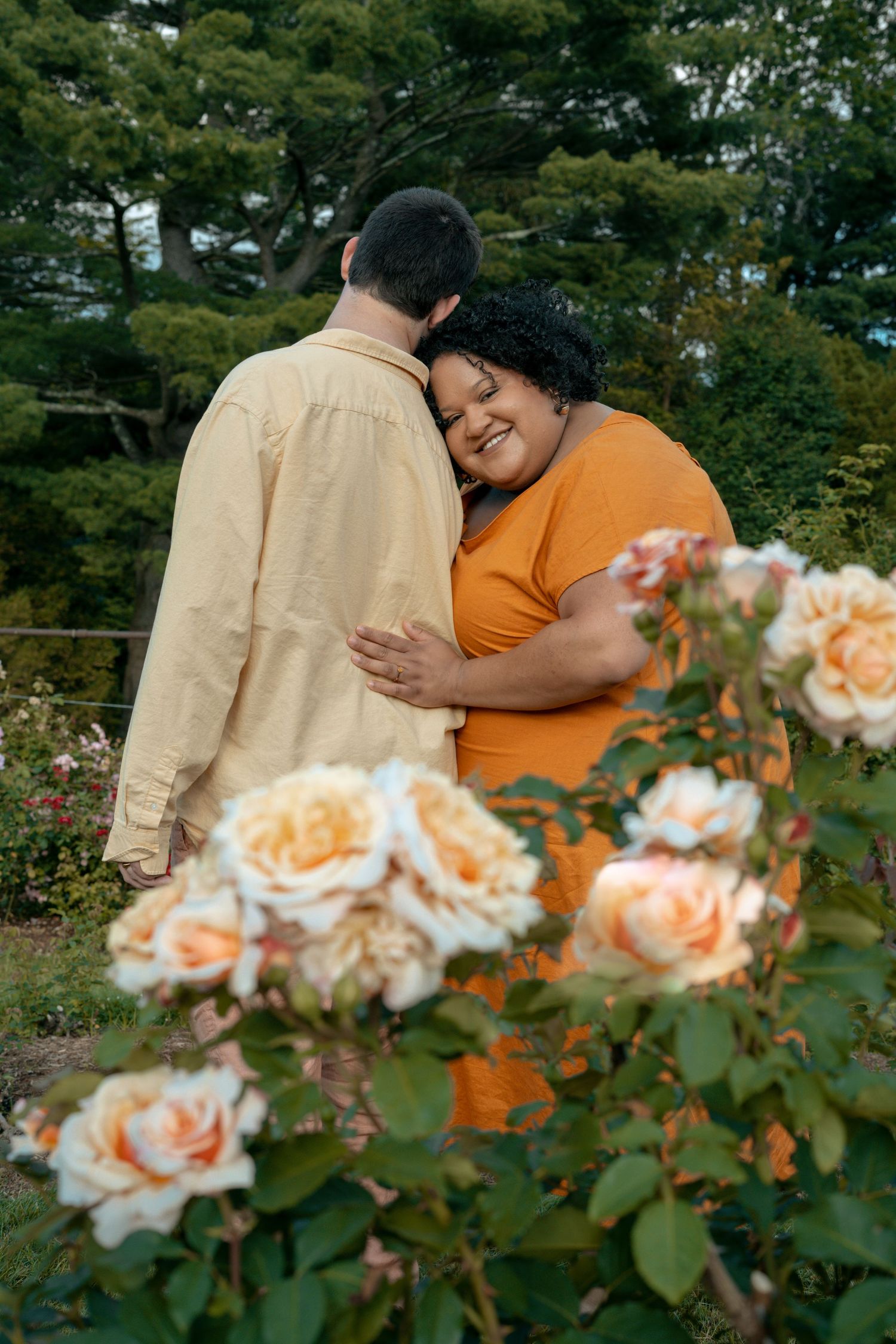 A plus-size Black woman in an orange top embraces a man in a garden with blooming peach roses.