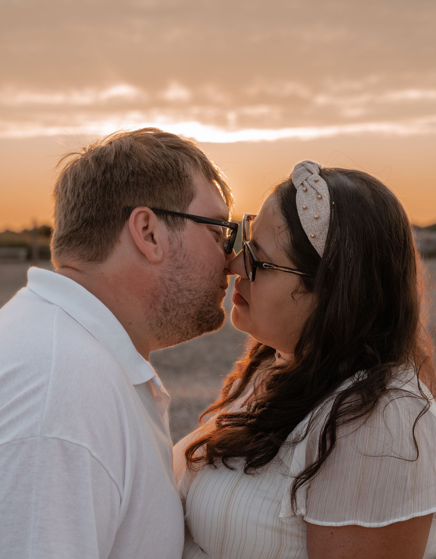 Couple sharing a kiss at sunset, both wearing sunglasses and white outfits outdoors.