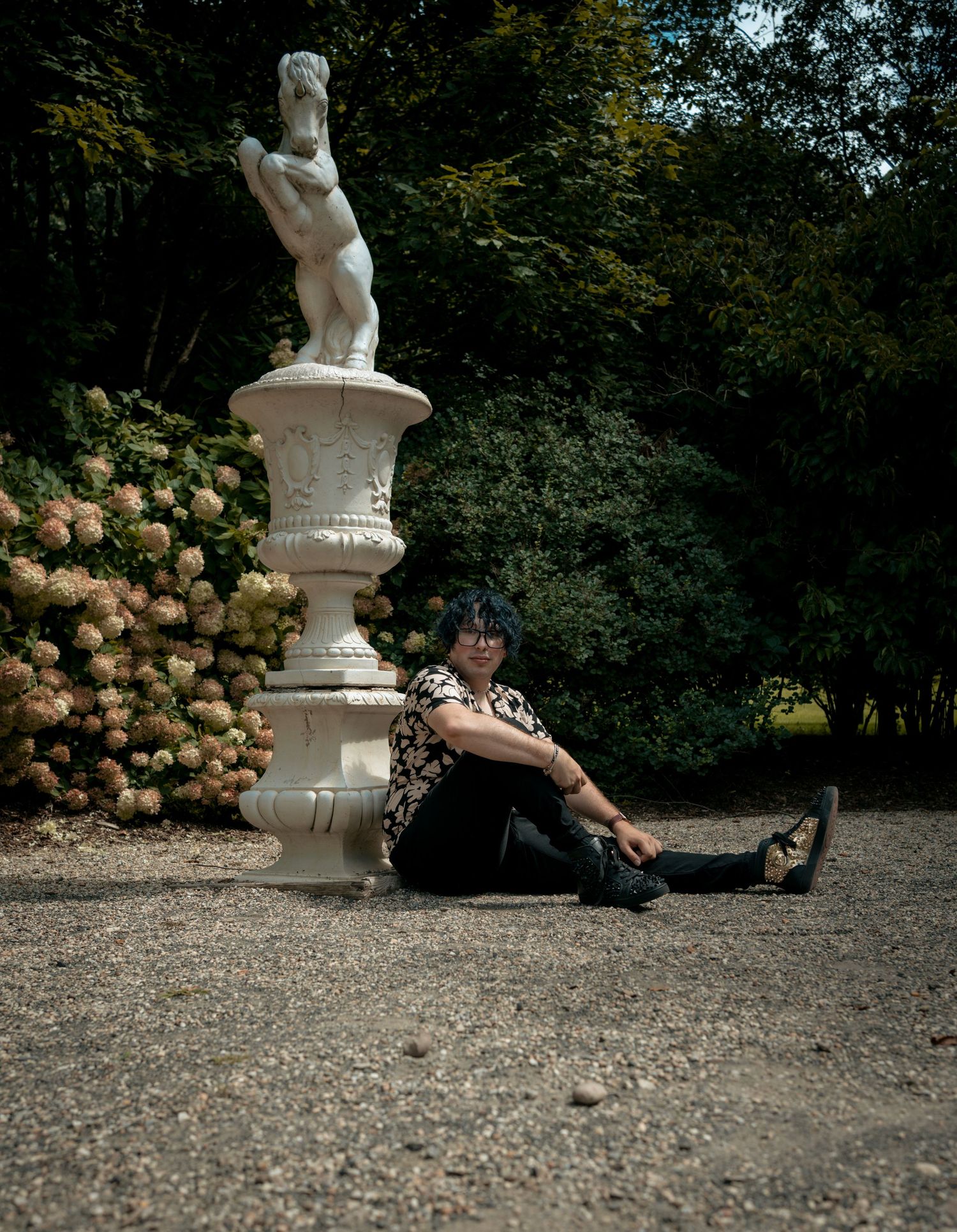 Person with blue hair sitting beside classical white statue in garden.