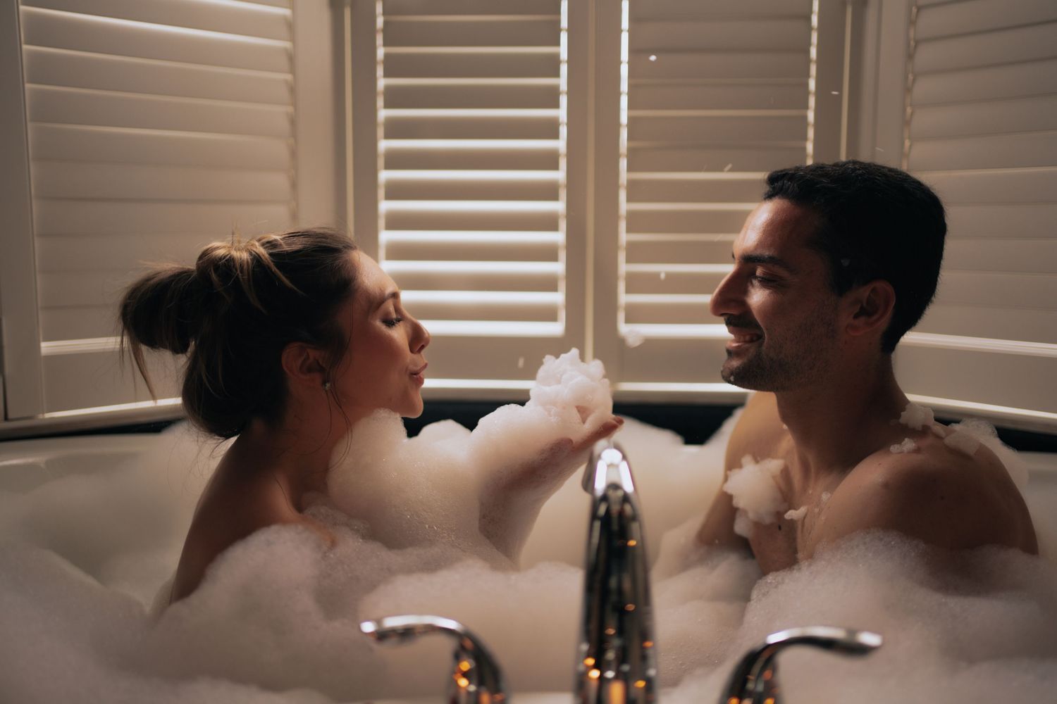 Couple enjoying a romantic bubble bath together near shuttered windows.