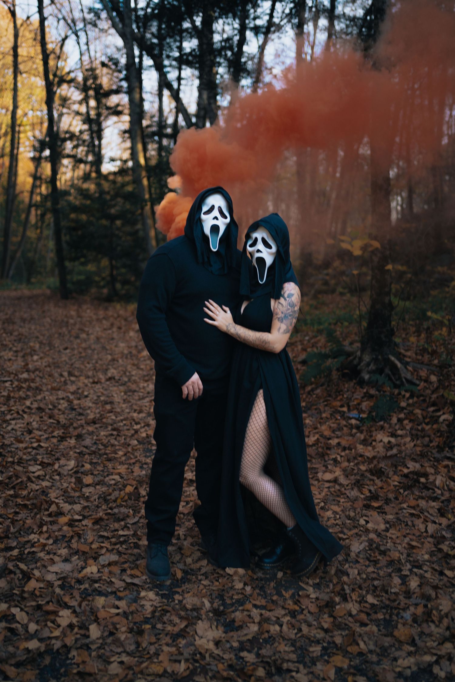 Couple in black Ghostface Scream costumes posing in autumn forest with orange smoke bomb.