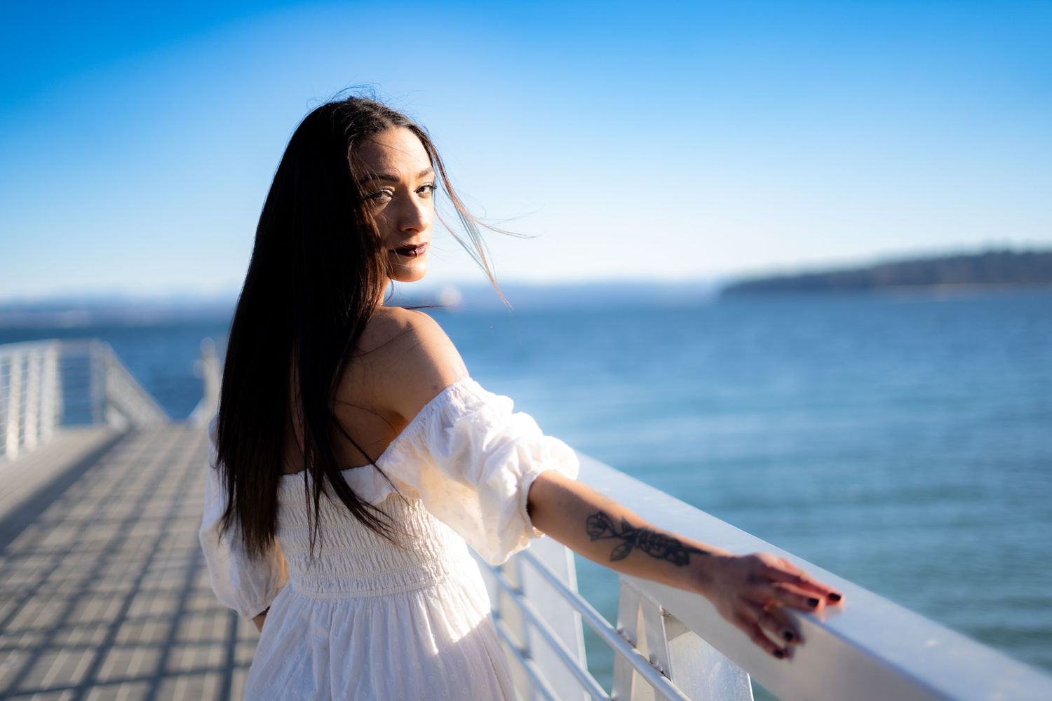 Woman in white off-shoulder dress leaning on pier railing with ocean backdrop.