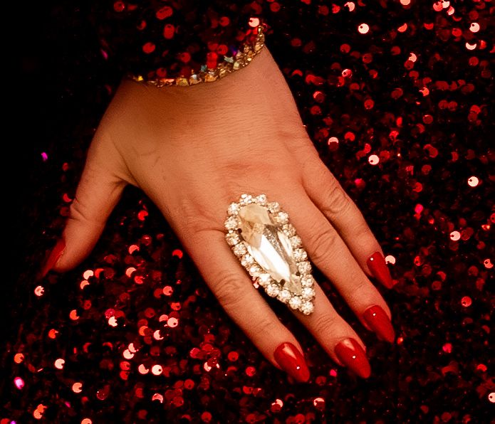 Hand wearing glamorous pearl and crystal statement ring with red manicure against sparkly red bokeh background.