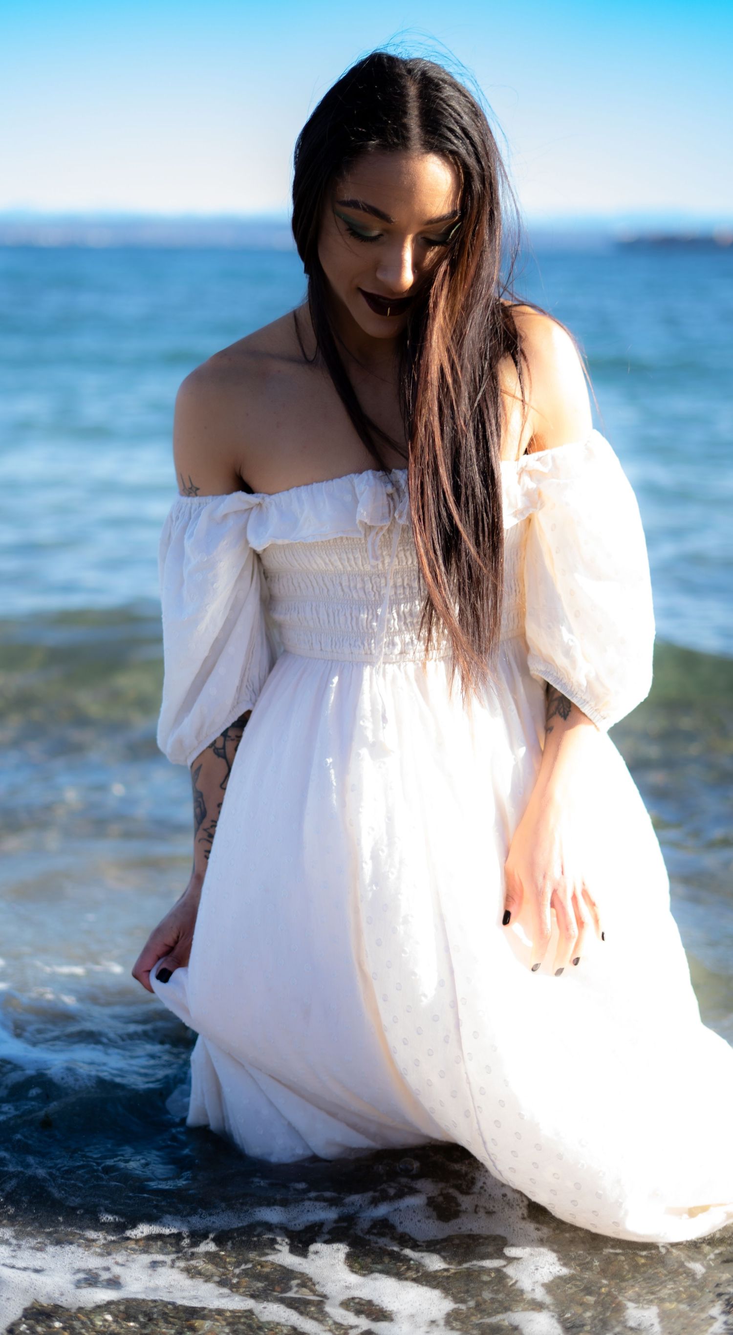 Woman in white off-shoulder dress standing in ocean waves on sunny beach.