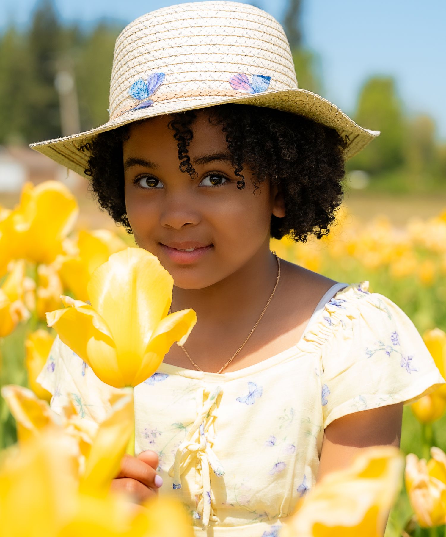 Close-up portrait of girl in straw hat holding yellow tulip in sunny flower field.