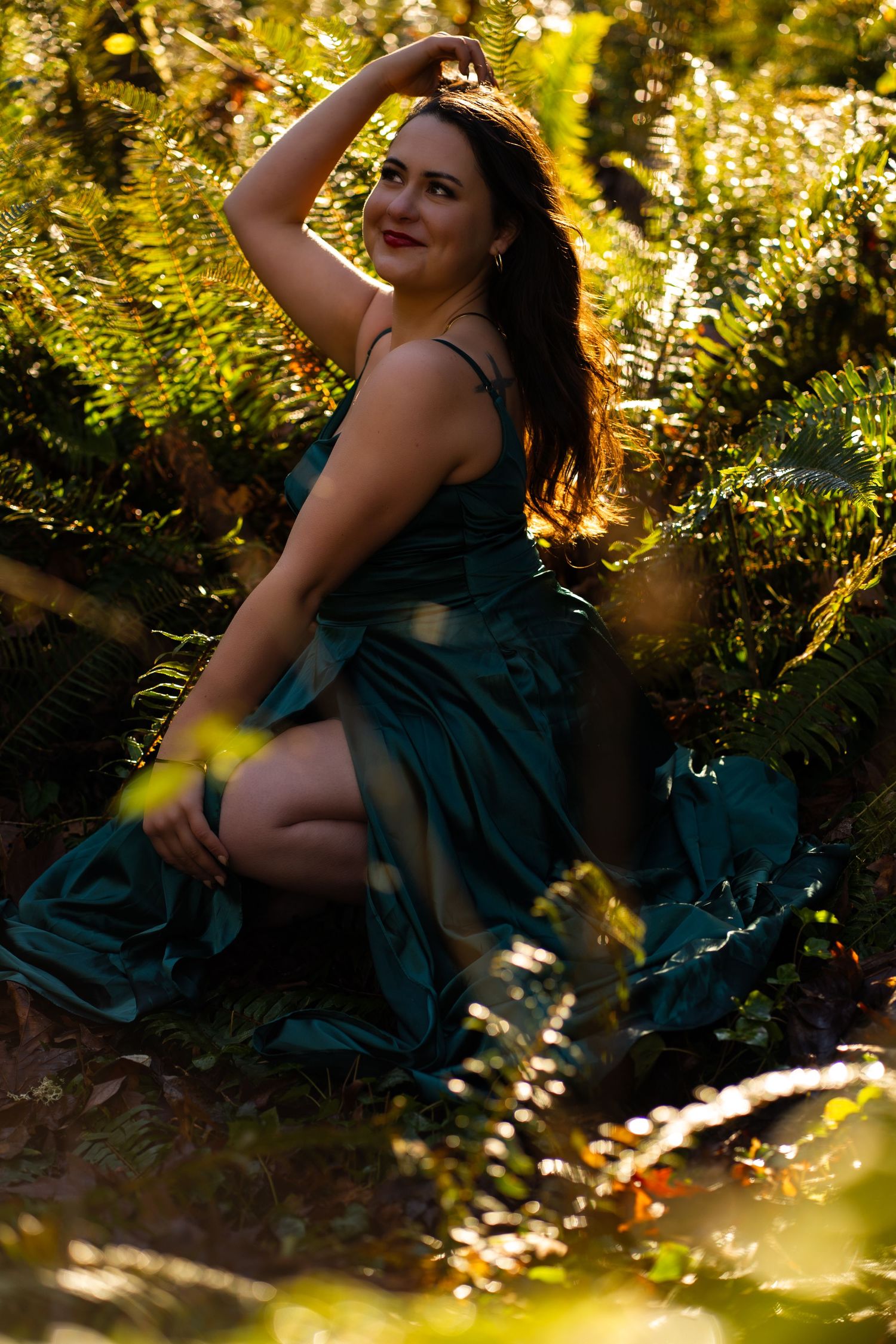 Woman in teal satin gown sitting gracefully among lush green forest ferns in sunlight.