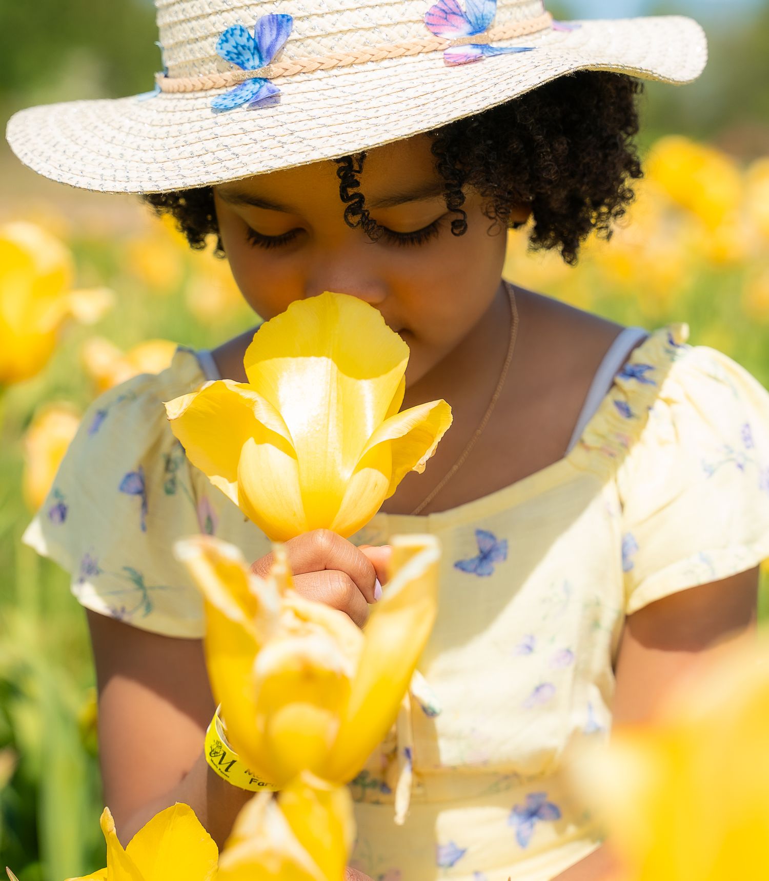 Young Black girl in yellow dress and butterfly hat smelling yellow tulips in flower field.