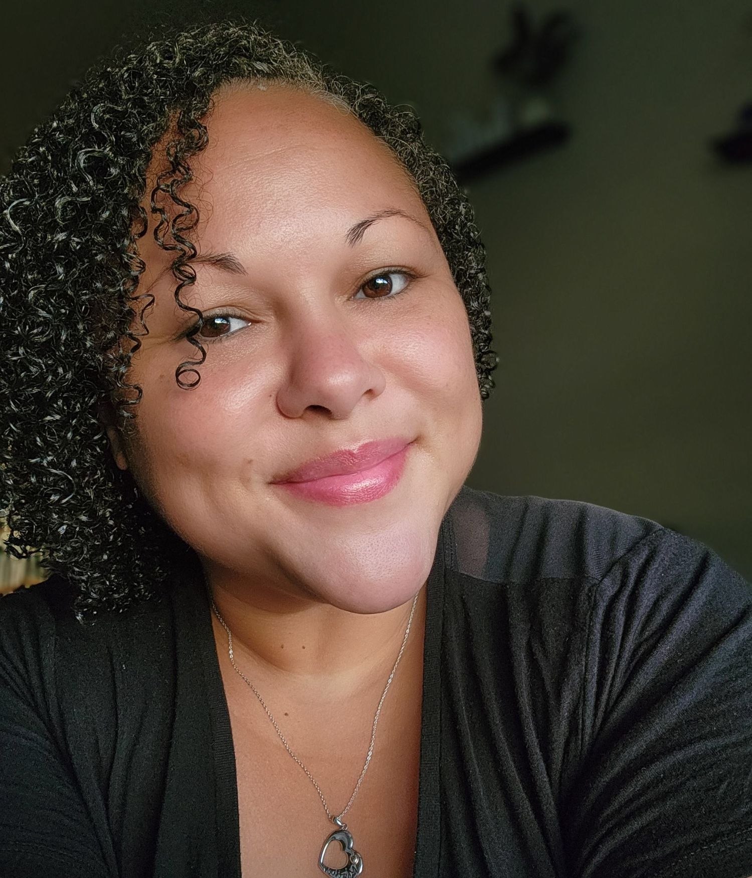 A smiling woman with natural curly hair wearing a black V-neck top and delicate silver necklace in a portrait photo.