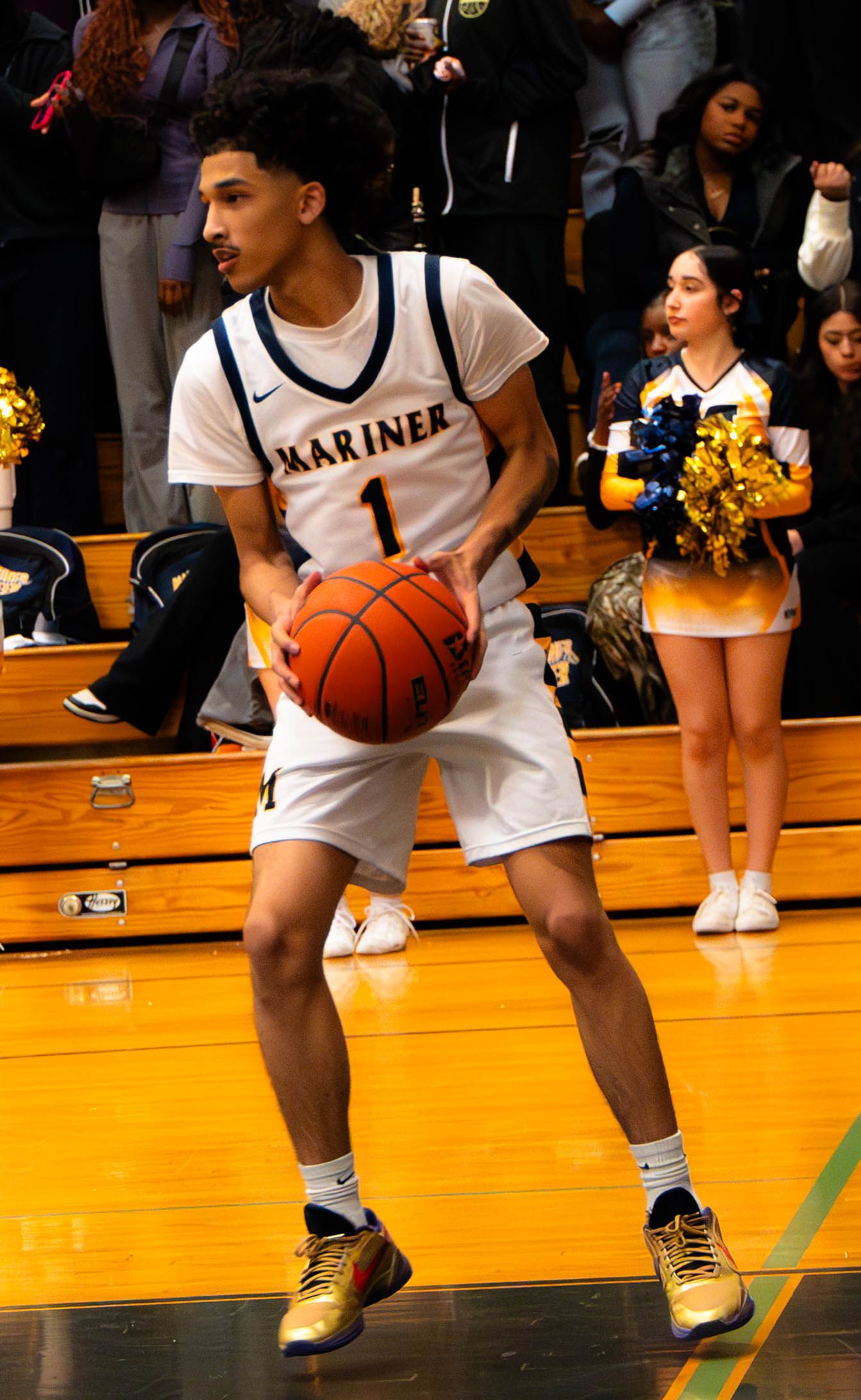 Mariner High School basketball player number 1 holds ball on court with cheerleaders visible in background.