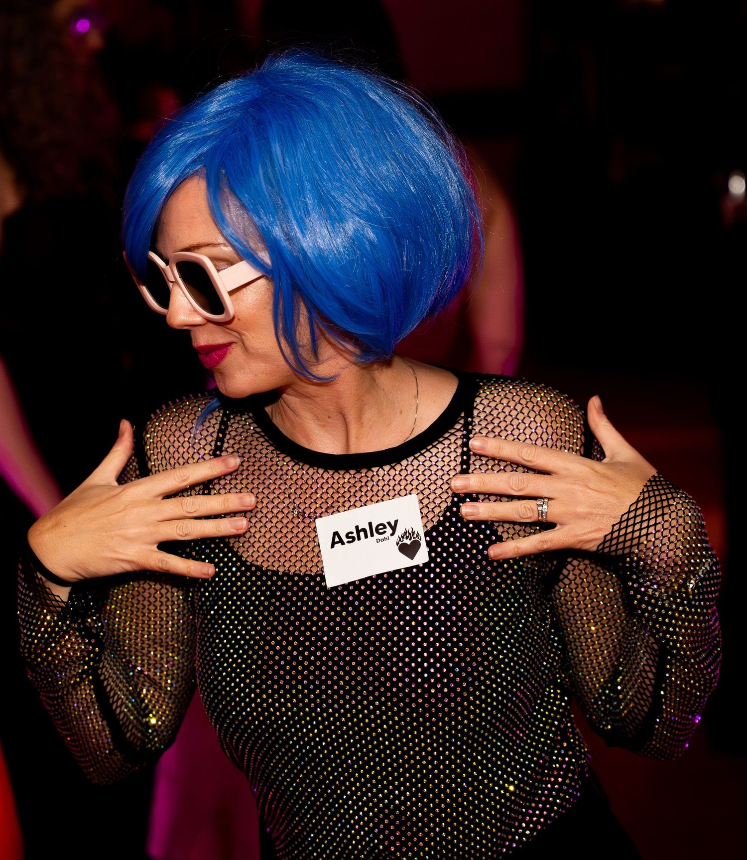 Woman in blue bob wig and mesh outfit wearing Ashley name tag dances at party.
