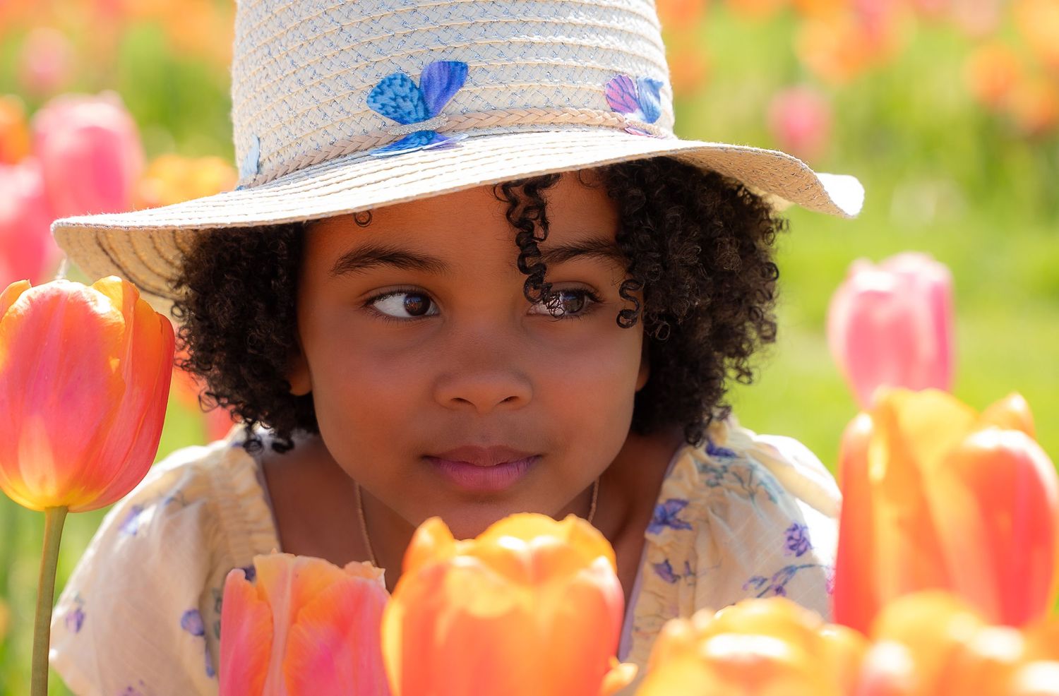 Young girl in straw hat surrounded by colorful pink and orange tulips in a field.