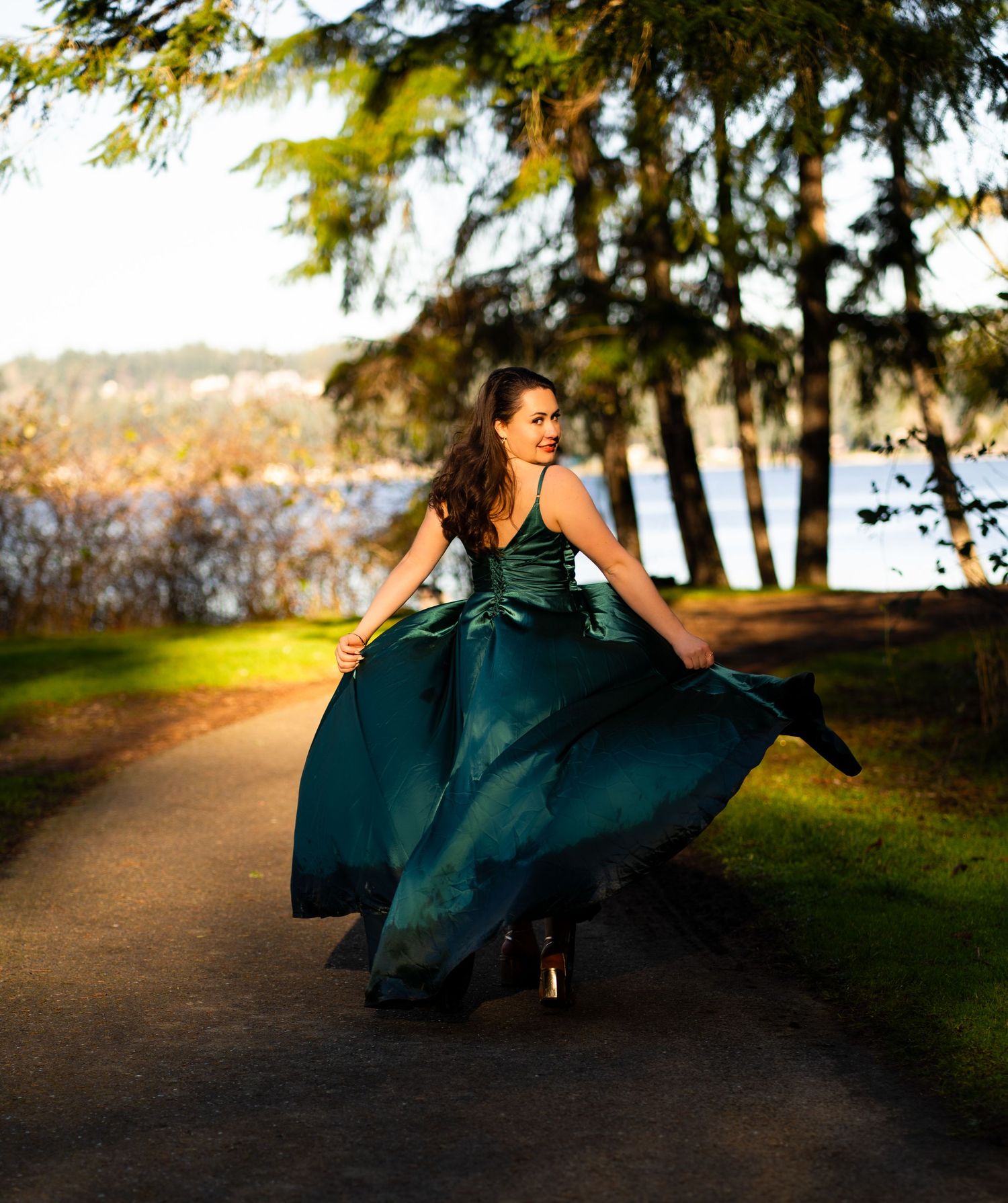 Woman in flowing green ball gown twirling on a tree-lined path at golden hour by a lake.