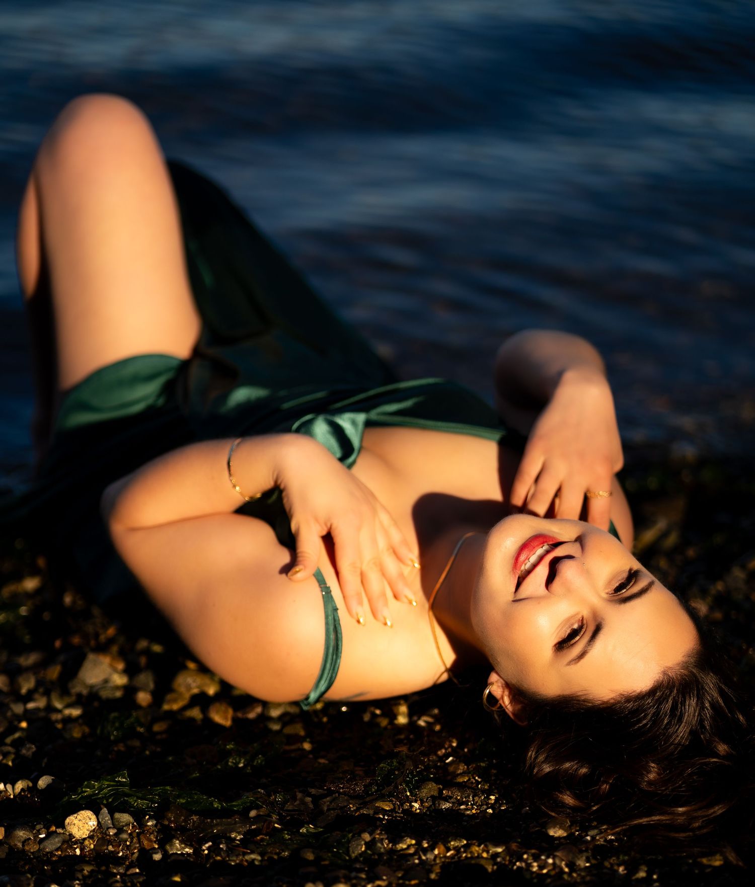 Woman in green satin dress lying on rocky shoreline beside glistening water.