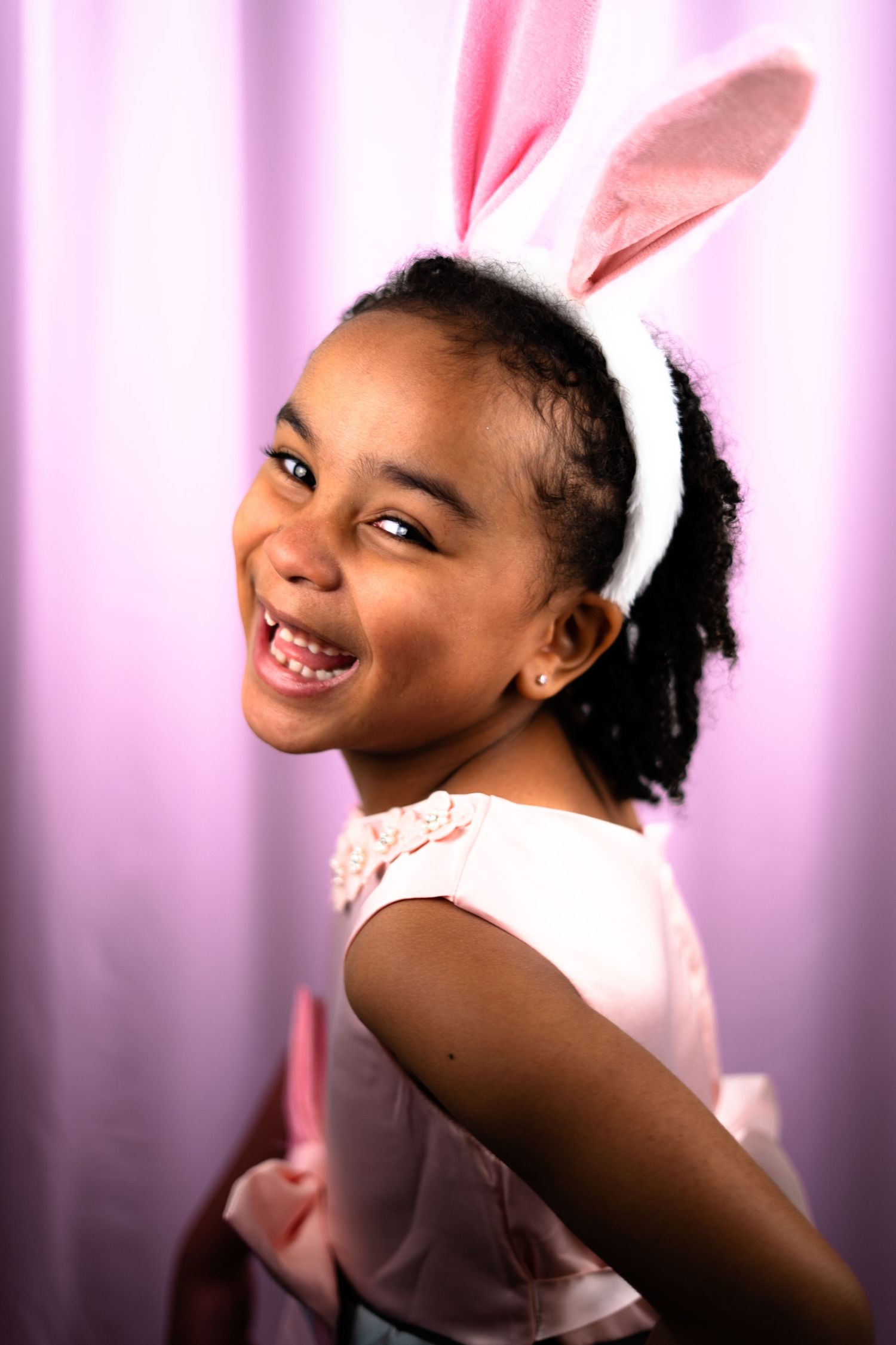 Joyful young girl wearing pink Easter bunny ears laughing against a soft pink curtain background.
