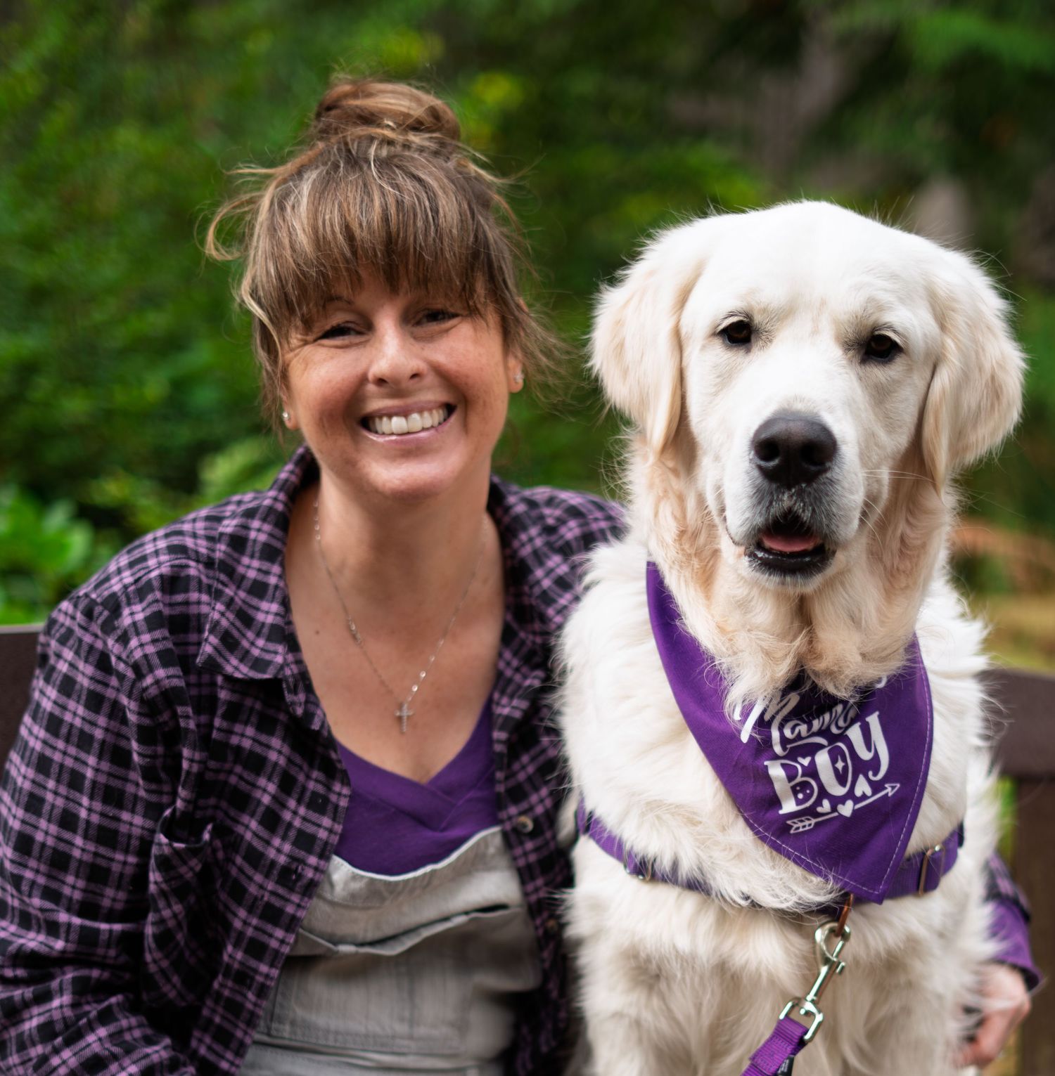 A smiling woman in a purple shirt poses with a white Golden Retriever wearing a purple bandana outdoors.