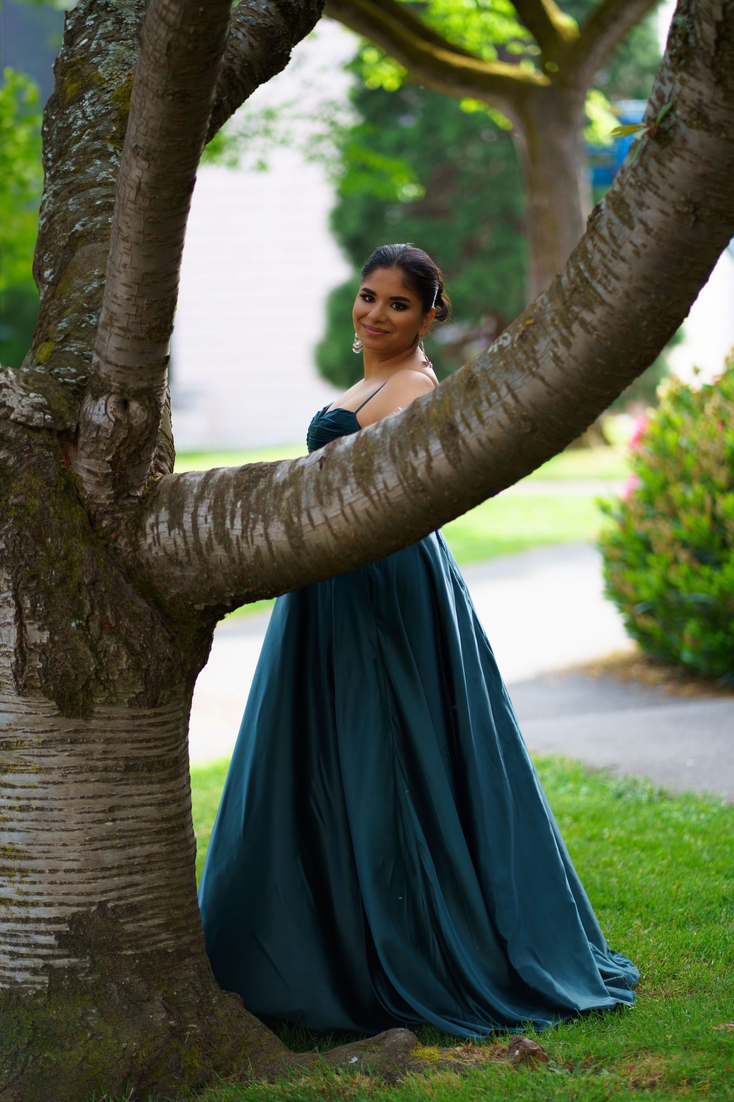 Woman in teal ball gown smiling while leaning against curved tree trunk in park.