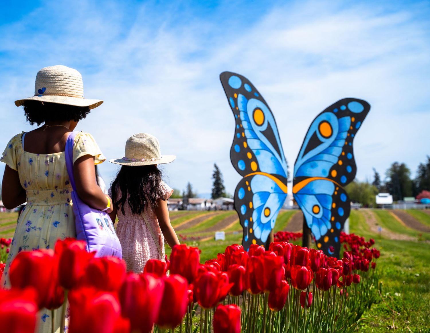 Two girls in straw hats walking past red tulips toward a large blue butterfly sculpture.