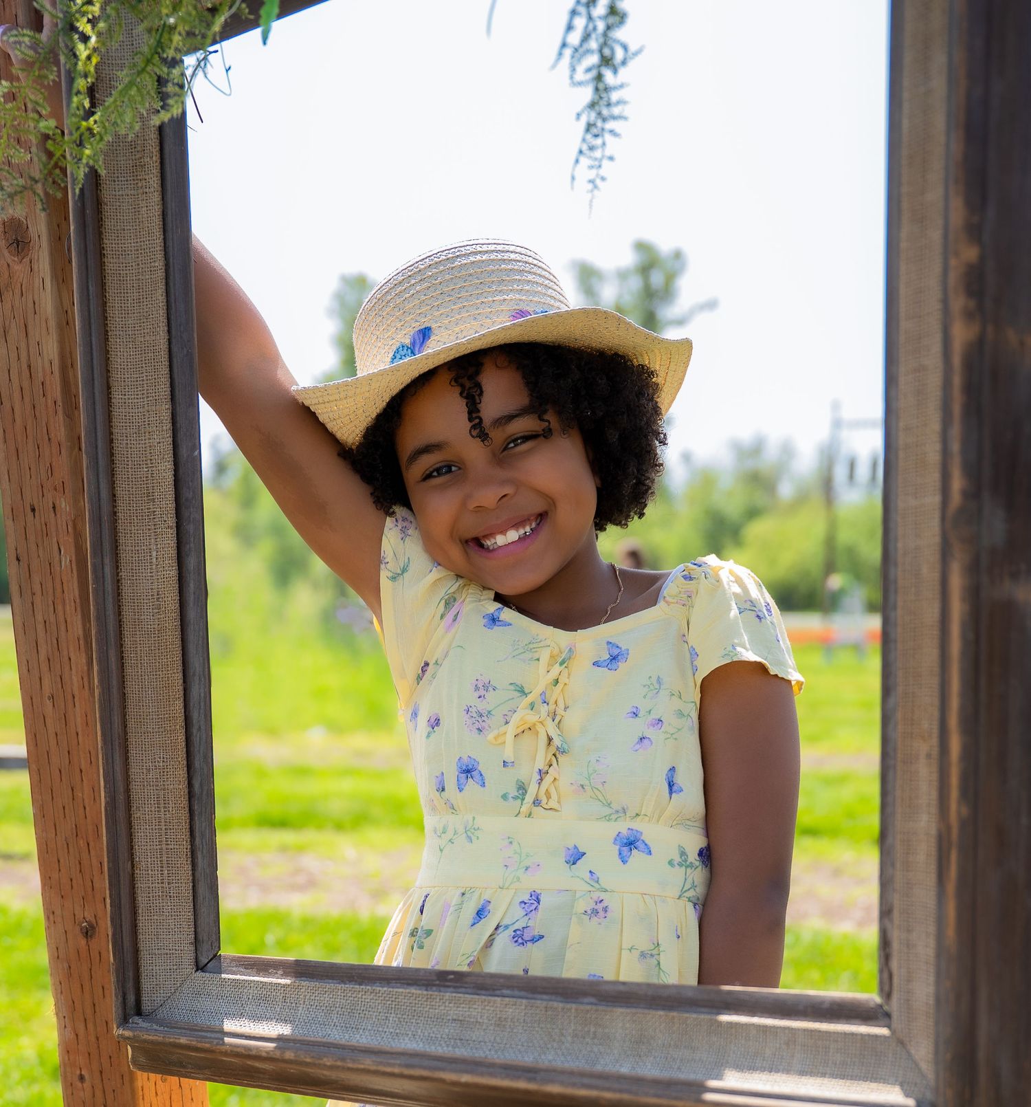 Smiling young girl in yellow floral dress and straw hat posing in a wooden frame outdoors.