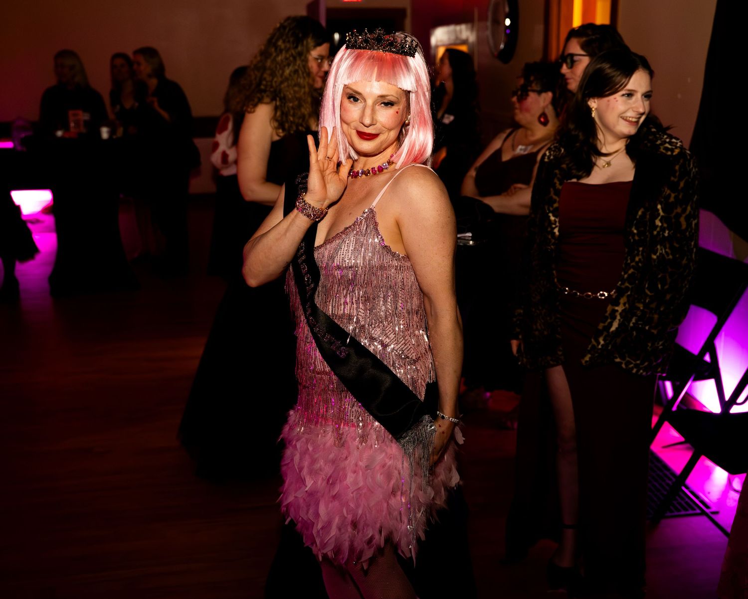 Performer in pink feathered dress and sash dances energetically on wooden floor during celebration event.