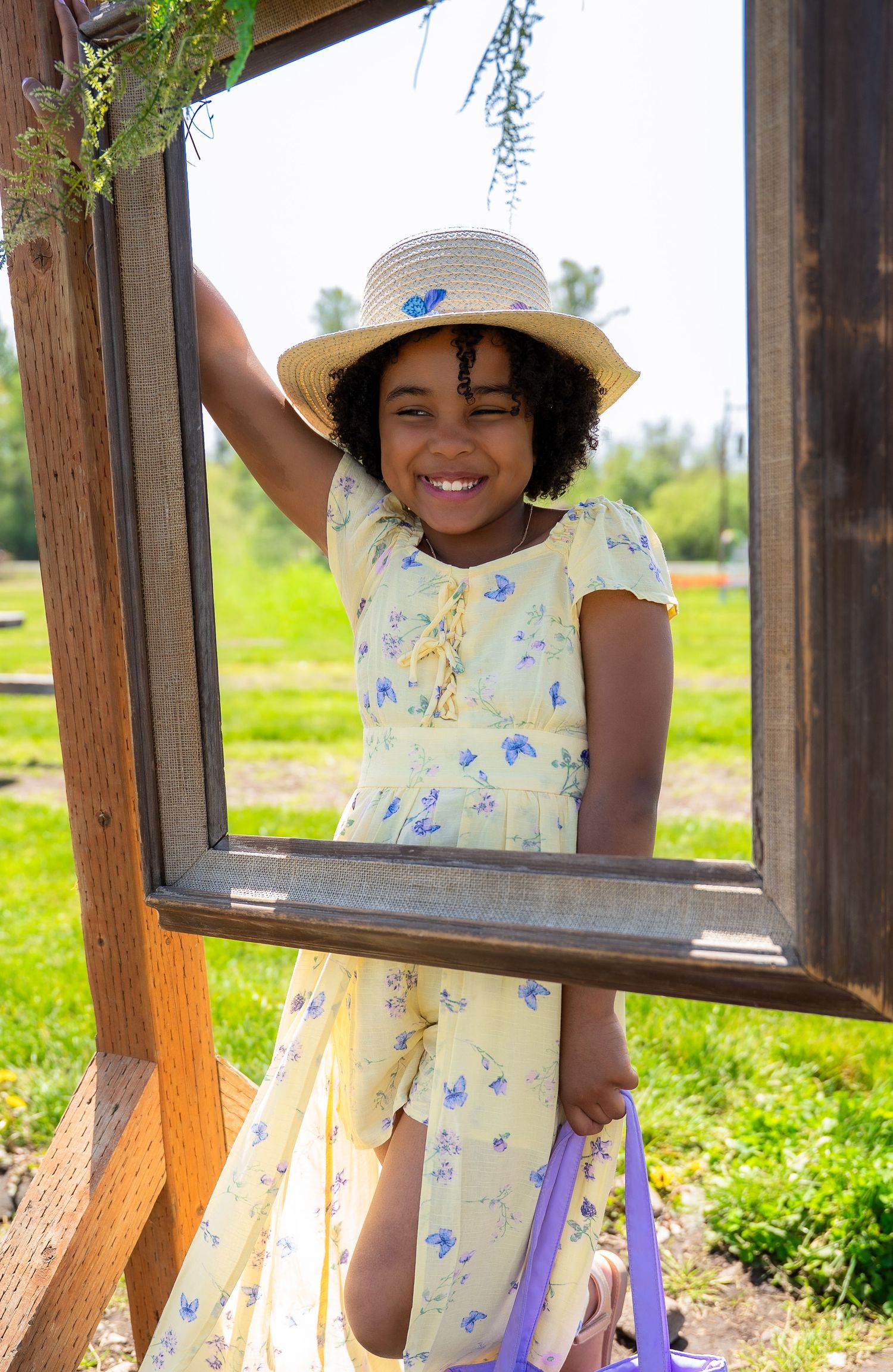 Smiling young girl in yellow floral dress and straw hat posing in a wooden frame outdoors.