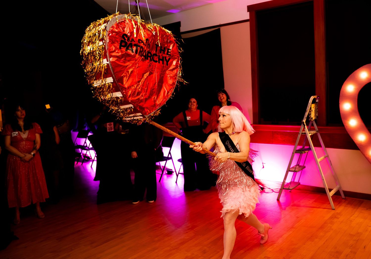 A performer in a pink fringe dress swings a bat at a gold heart-shaped pinata at a party.