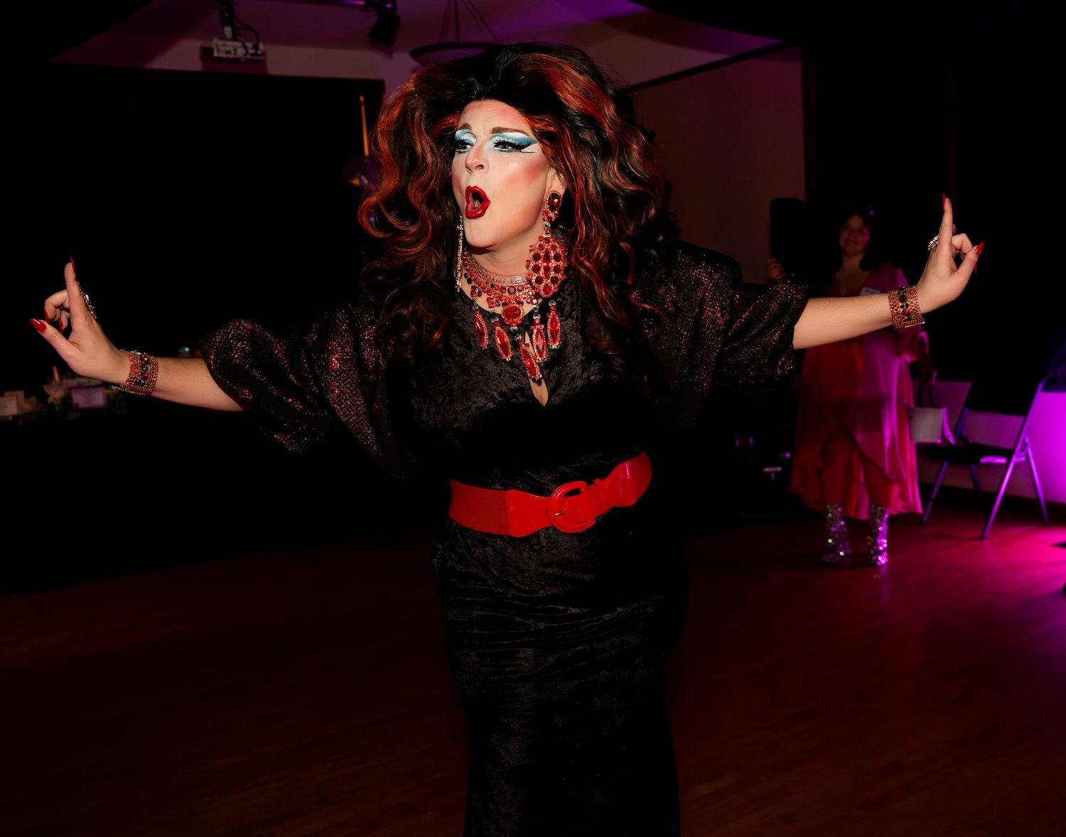 A drag queen in a black lace dress with red belt and beaded necklace performs on a dance floor.