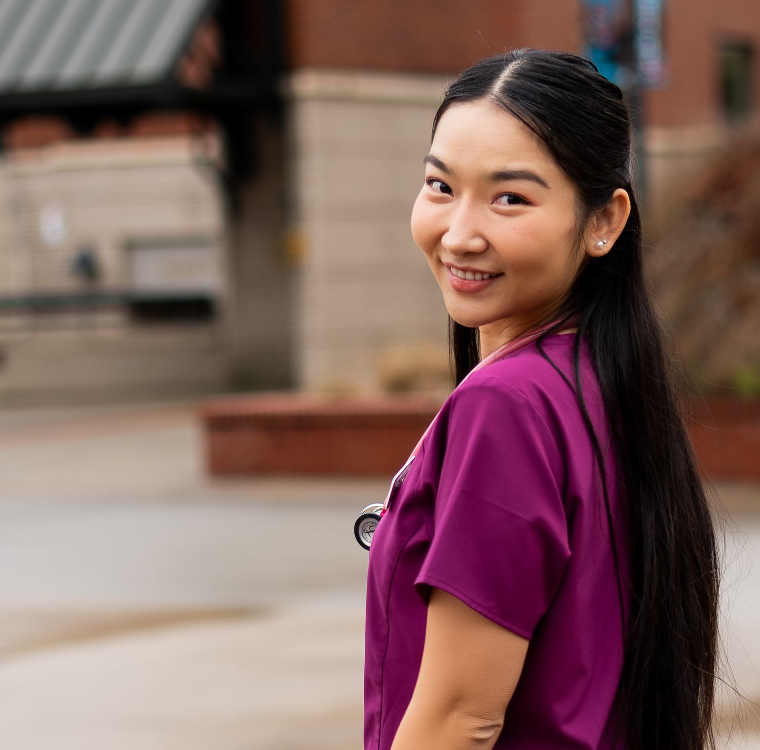 Smiling woman in magenta nursing scrubs with stethoscope outdoors on campus.