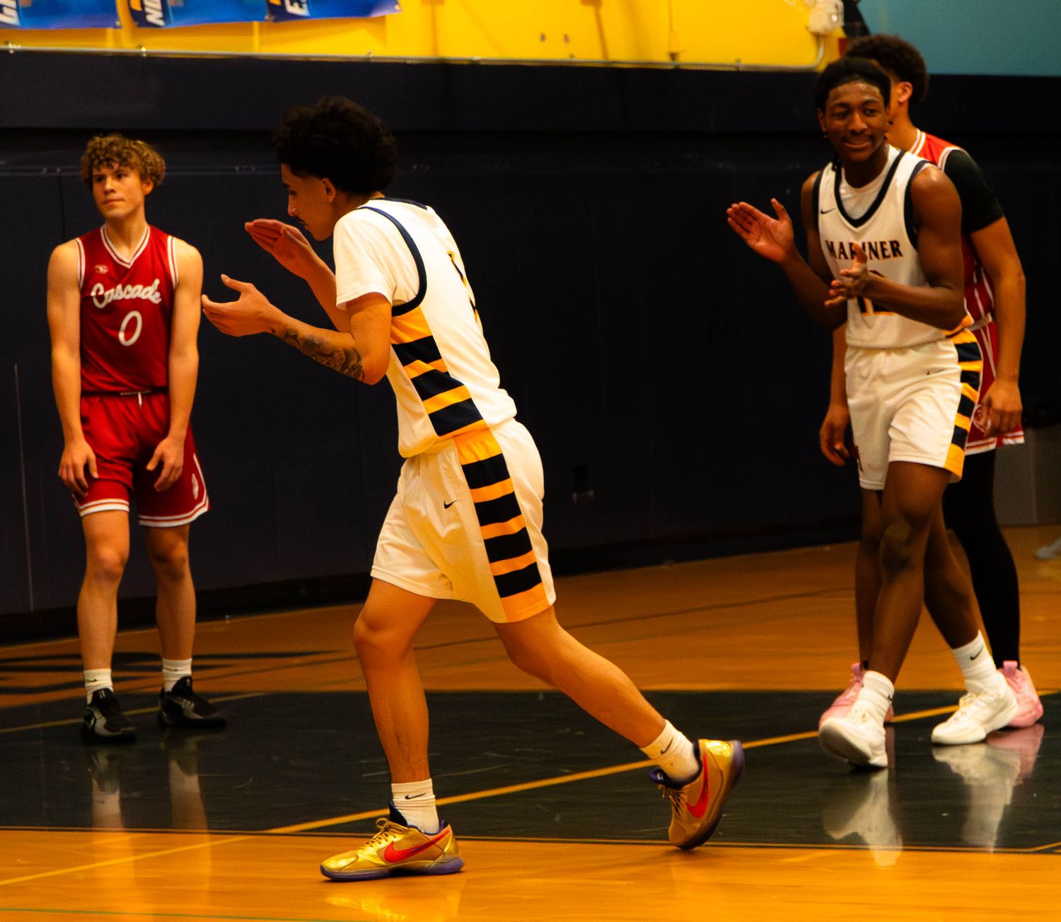 Two Mariner basketball players in white uniforms receive pass during home game against red-uniformed opponents.