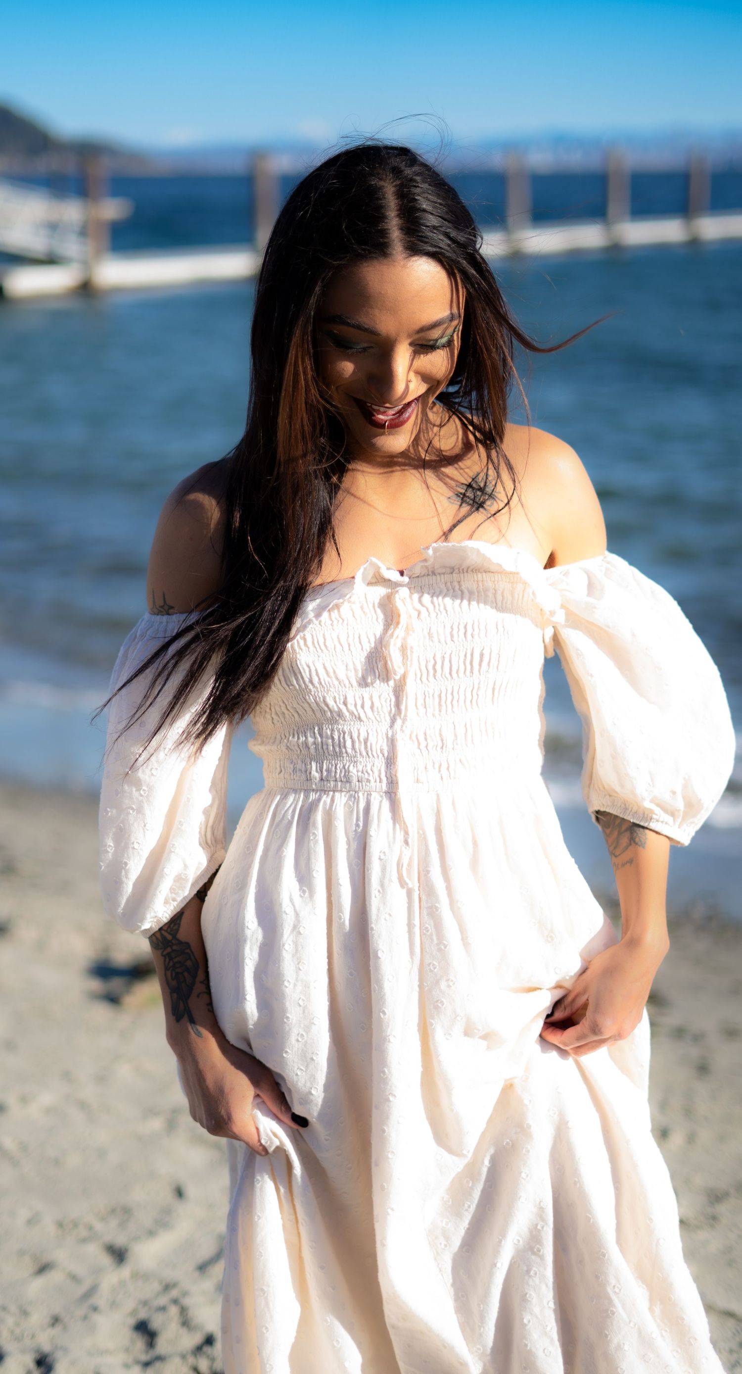 A smiling woman with long dark hair wears a white off-shoulder dress on a sunny beach by the ocean.