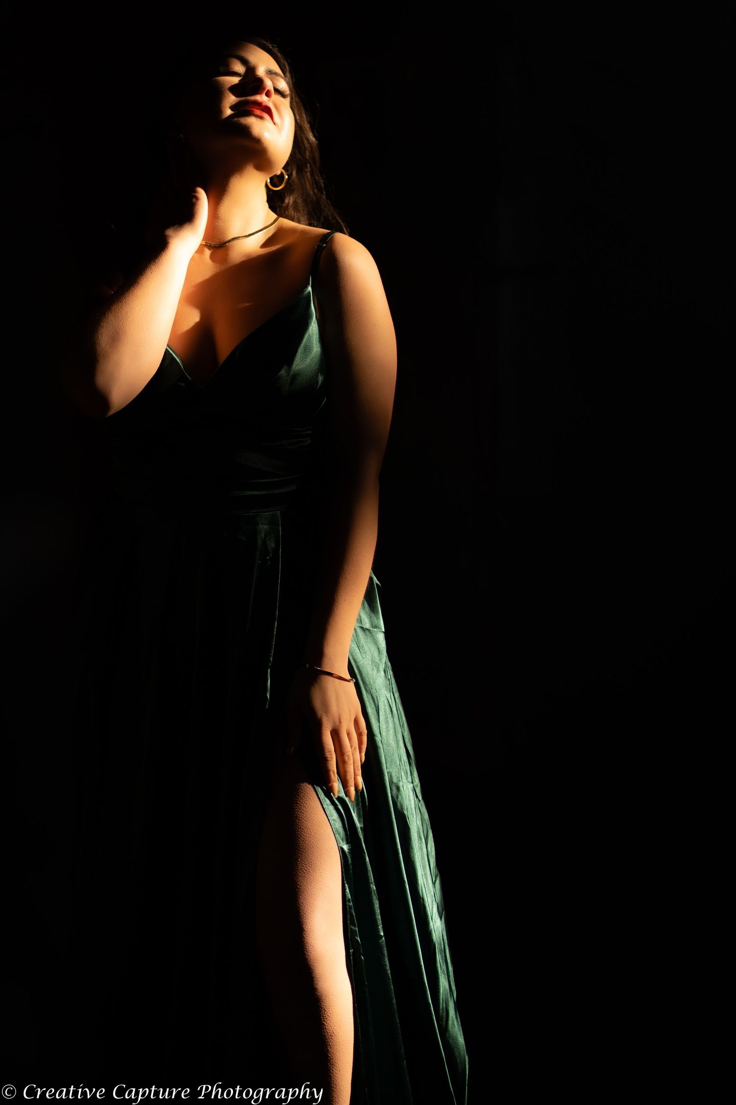 Woman in emerald green slit gown posing dramatically against black background in spotlight.
