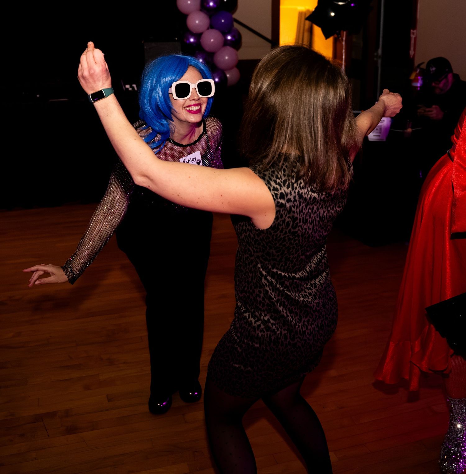 Two women dance together at a party, one wearing a blue wig and white sunglasses.
