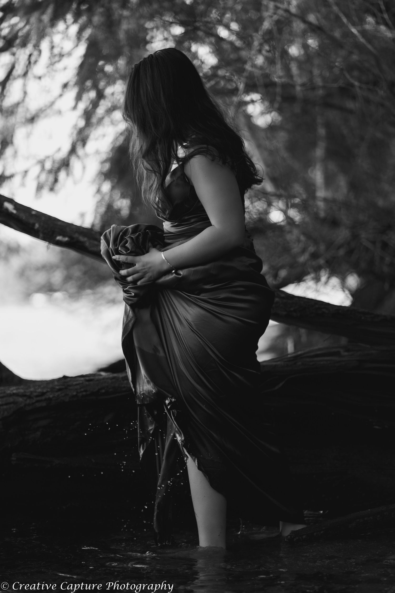 Black and white photo of woman in flowing dress standing in water near trees.
