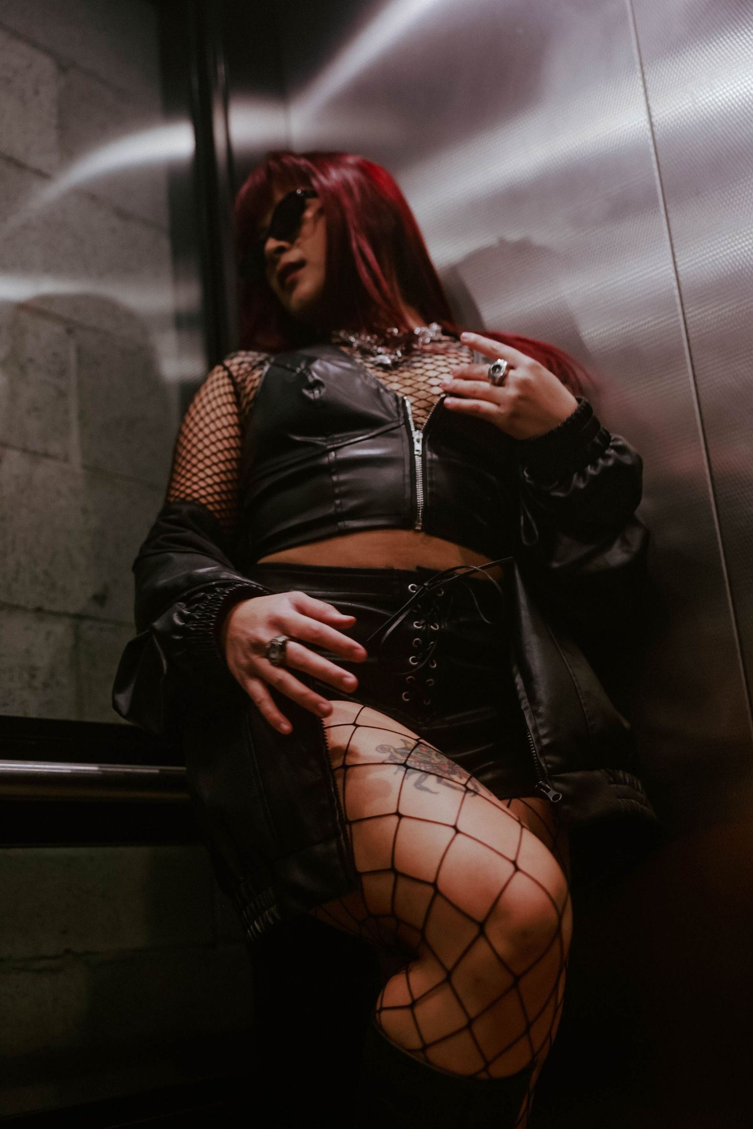 A red-haired woman in leather and fishnet outfit poses in a dimly lit elevator.