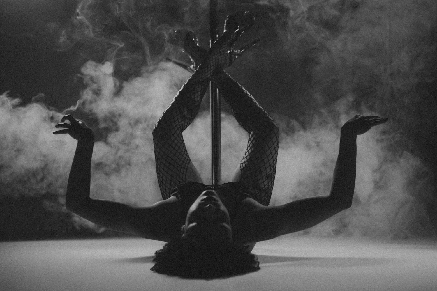 Black and white photo of a dancer performing an acrobatic pole dance move surrounded by dramatic smoke.