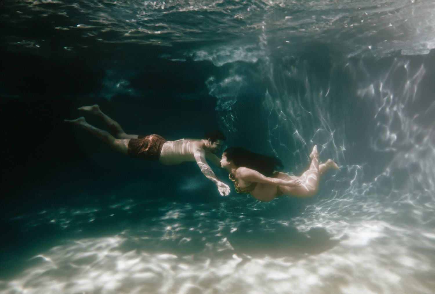 Two people swimming underwater in a clear blue pool, captured from below the surface.