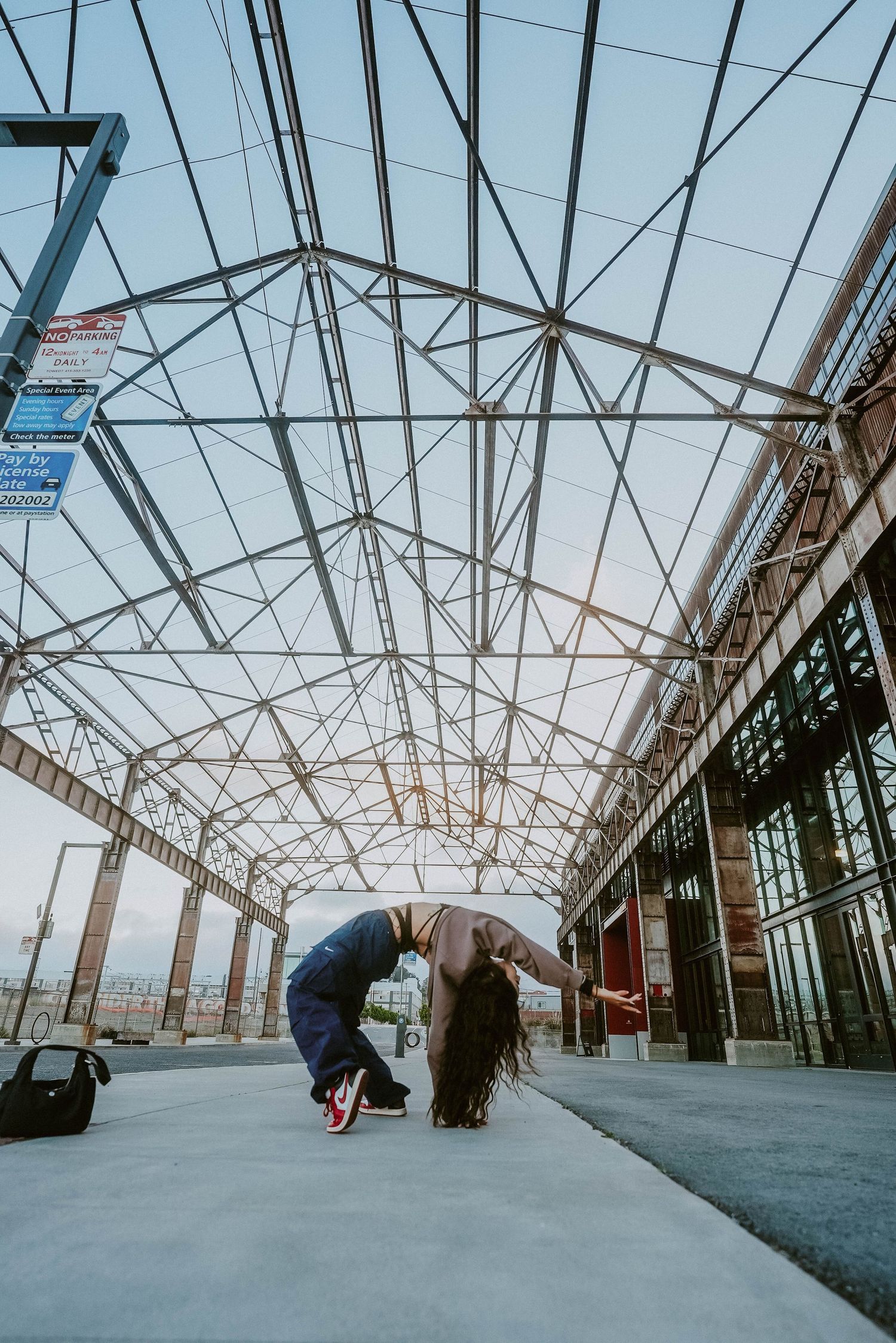 A breakdancer performs an impressive backbend move inside a large industrial building with a geometric steel roof structure.