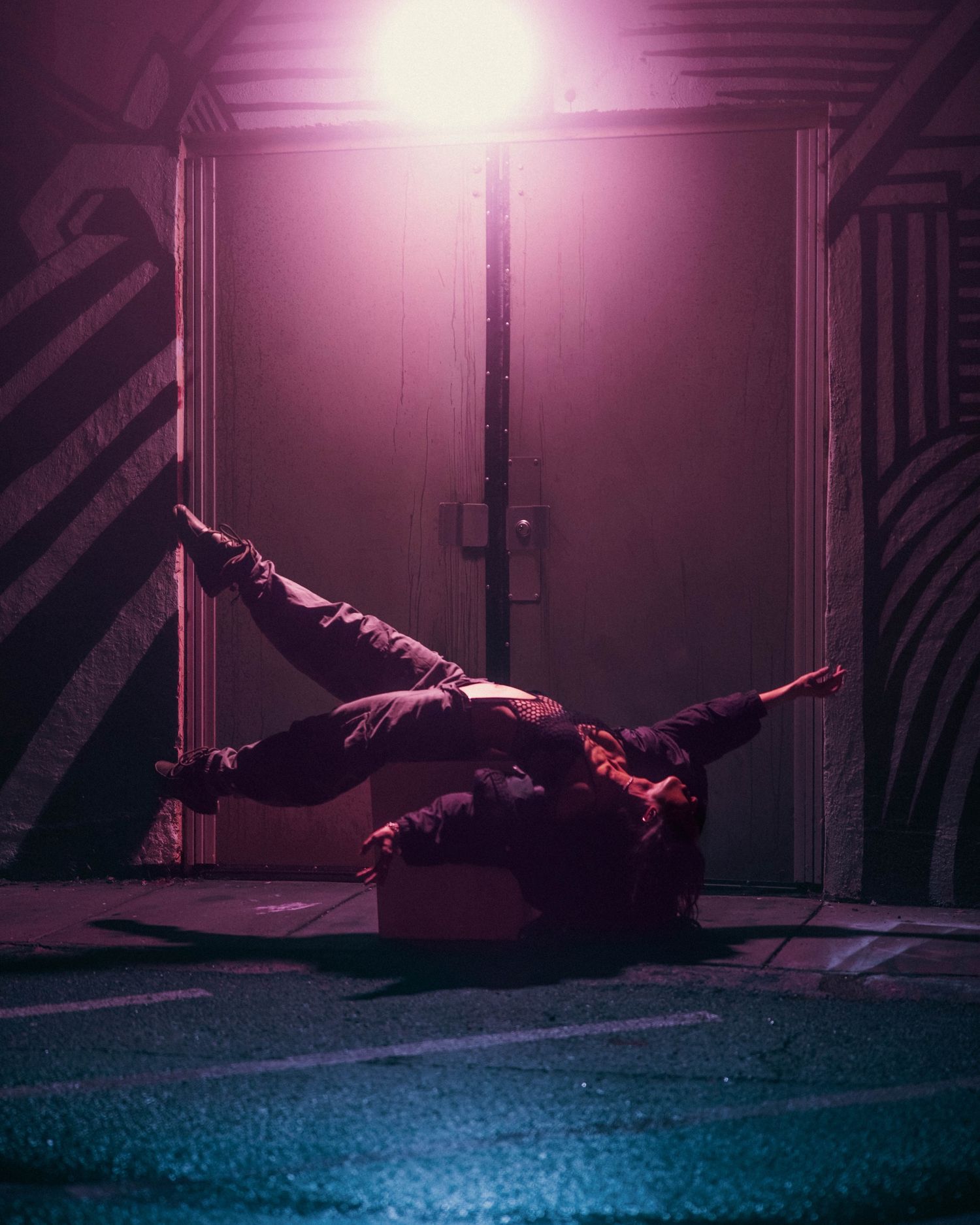 A performer balances horizontally under a glowing pink street lamp against a graffiti-covered wall at night.