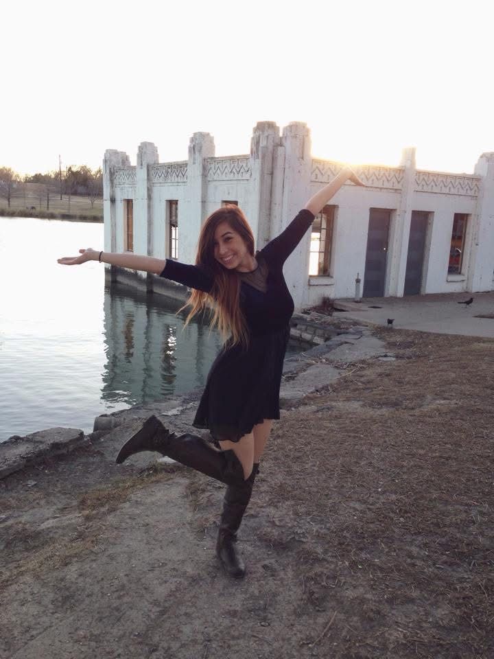 Woman in black dress and boots poses with arms outstretched near a white historic building by the water at sunset.