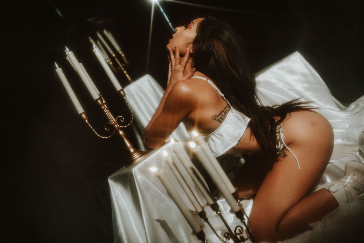 A woman in white lingerie poses dramatically beside a candelabra in moody dark lighting.