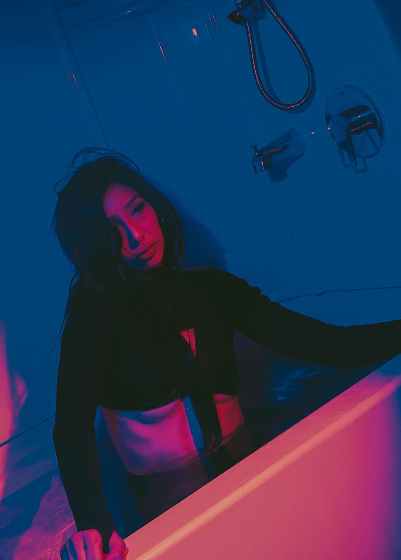 Young woman in black crop top poses by a bathtub in moody blue and pink neon lighting.