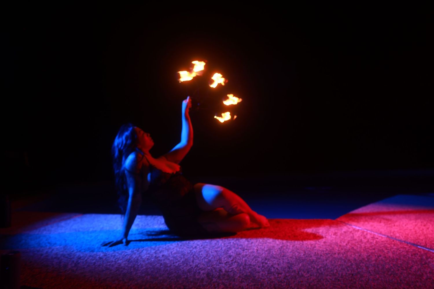 A fire dancer performs on a dark stage, lying gracefully while holding multiple flames against blue and red lighting.