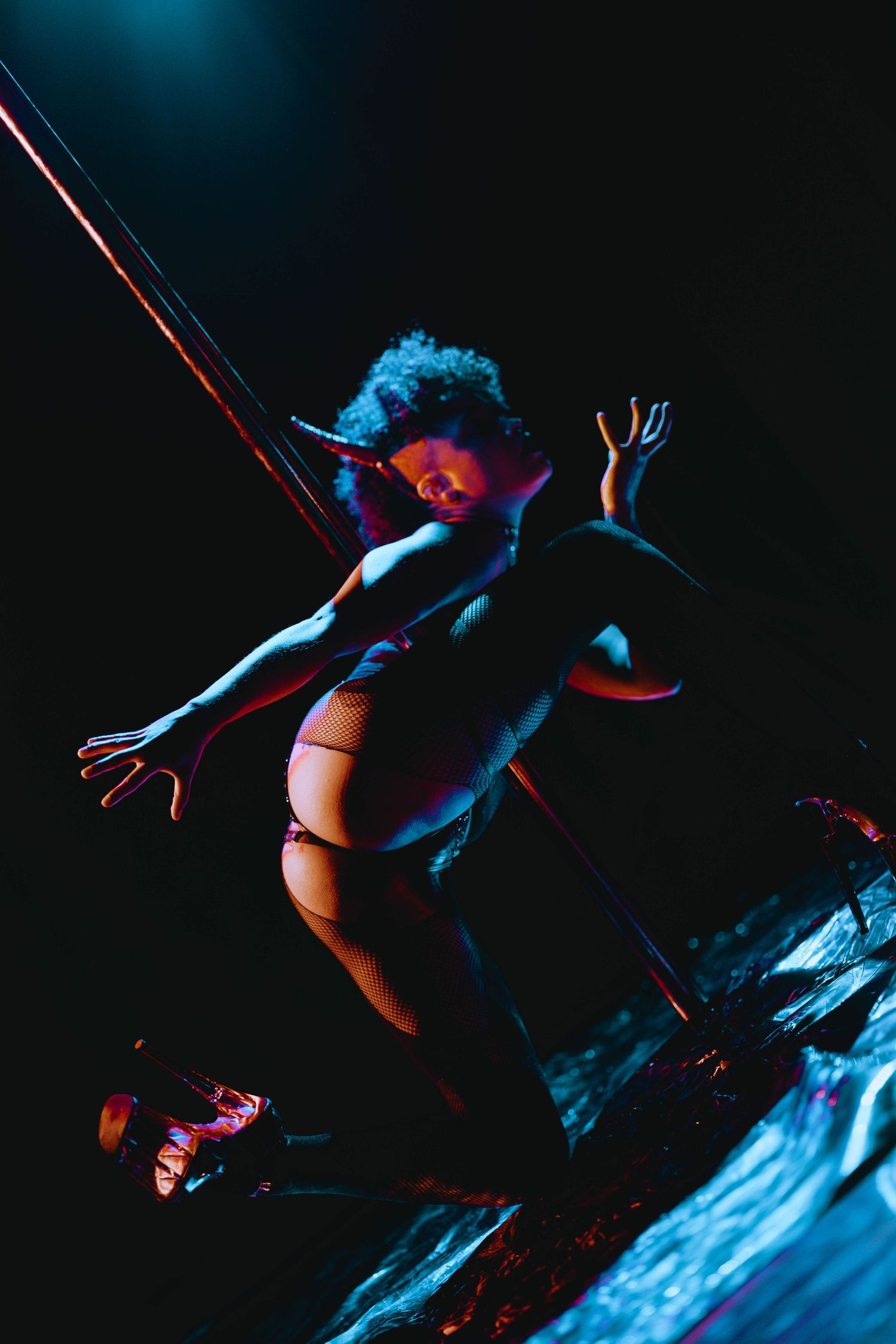 A pole dancer leaps gracefully near a pole under moody blue and purple stage lighting.