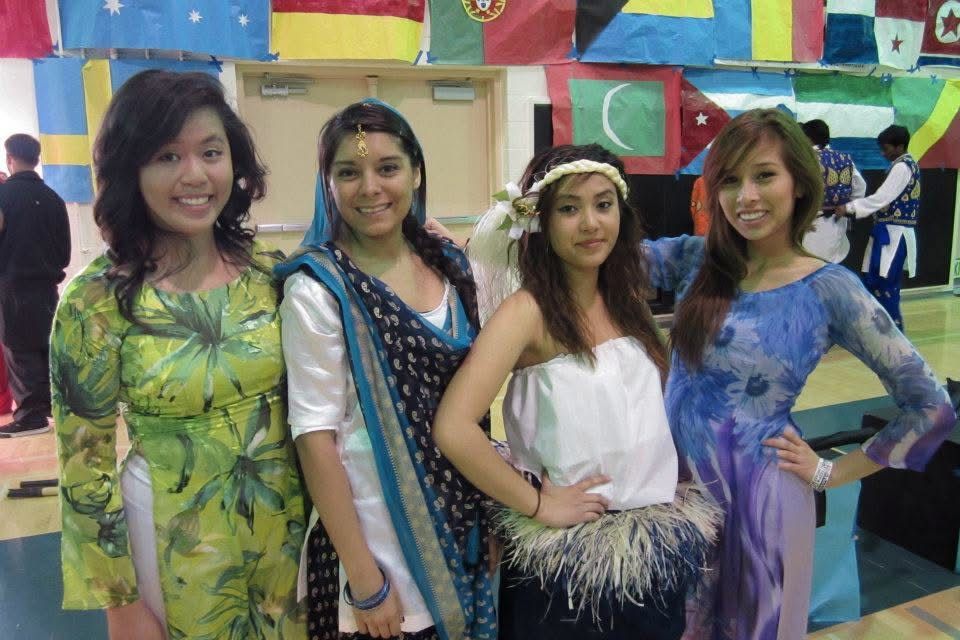 Four women in colorful traditional cultural costumes pose together at an international multicultural event with flags in background.
