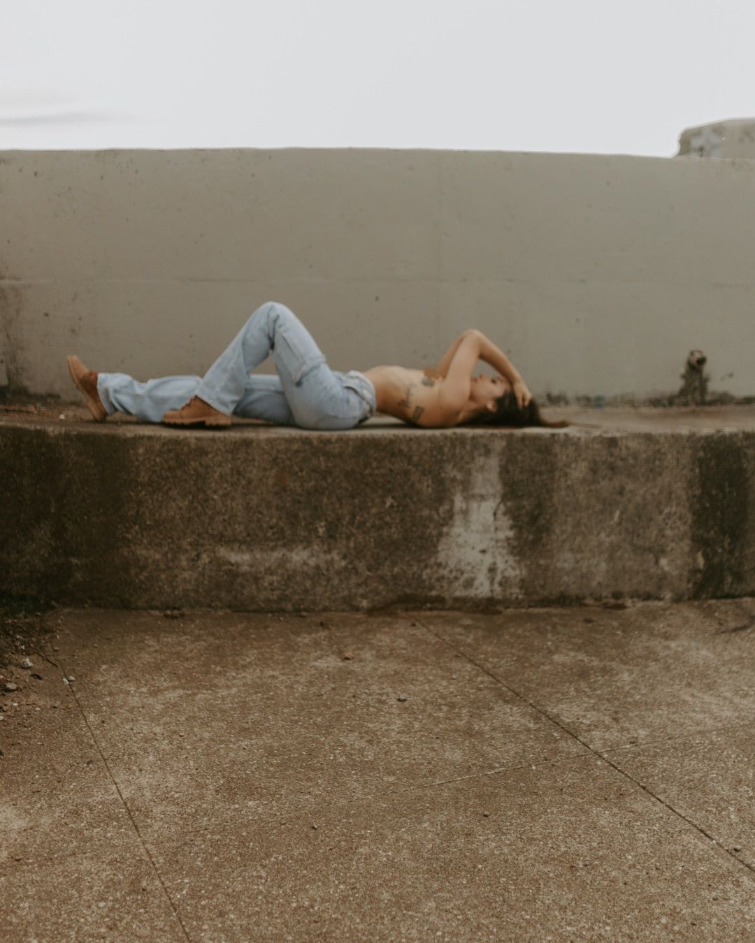 Person in light blue jeans lying on a concrete ledge outdoors against a beige wall.
